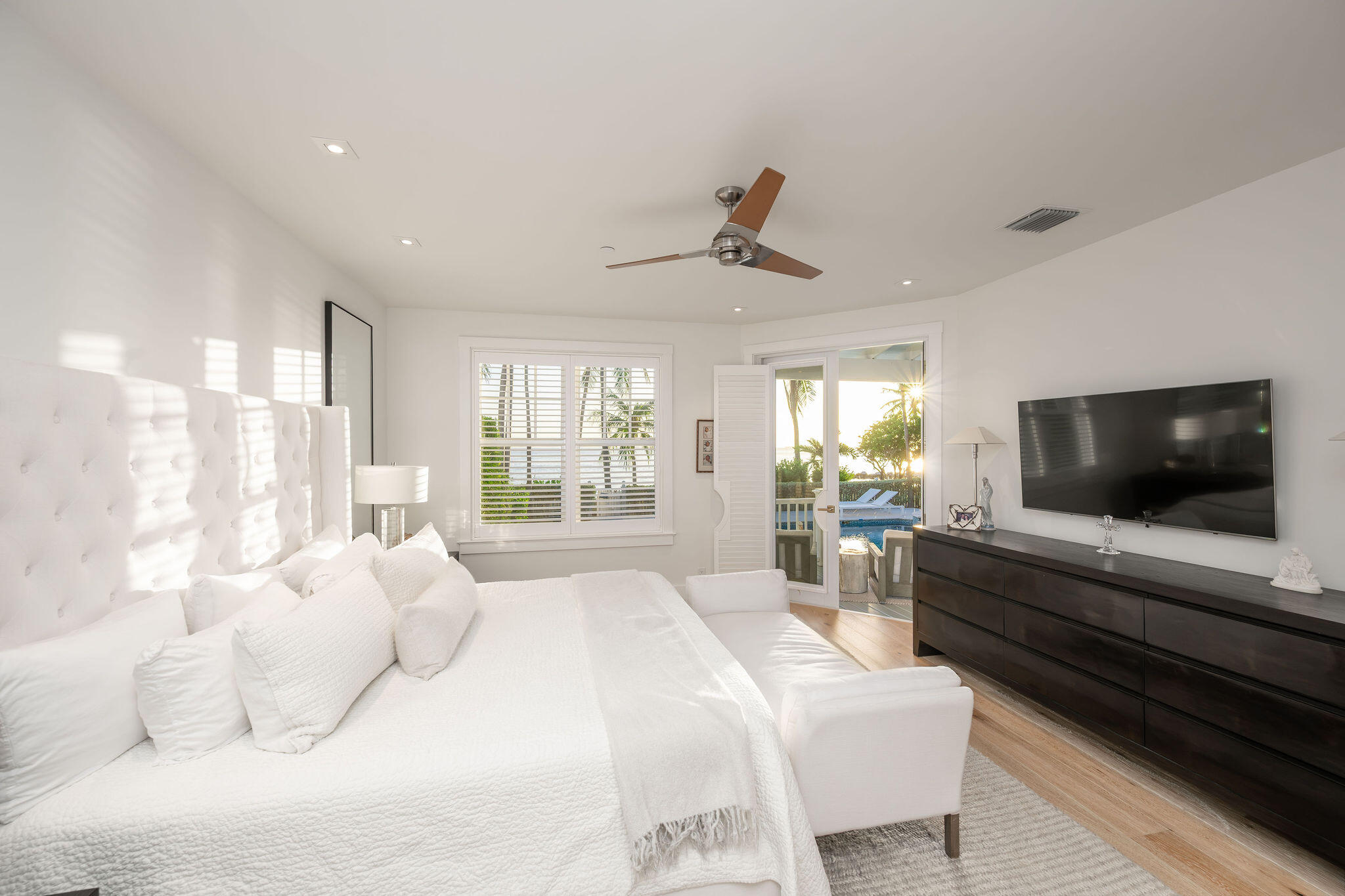 White bedroom with a large tufted bed and open doors overlooking a pool and palm trees.