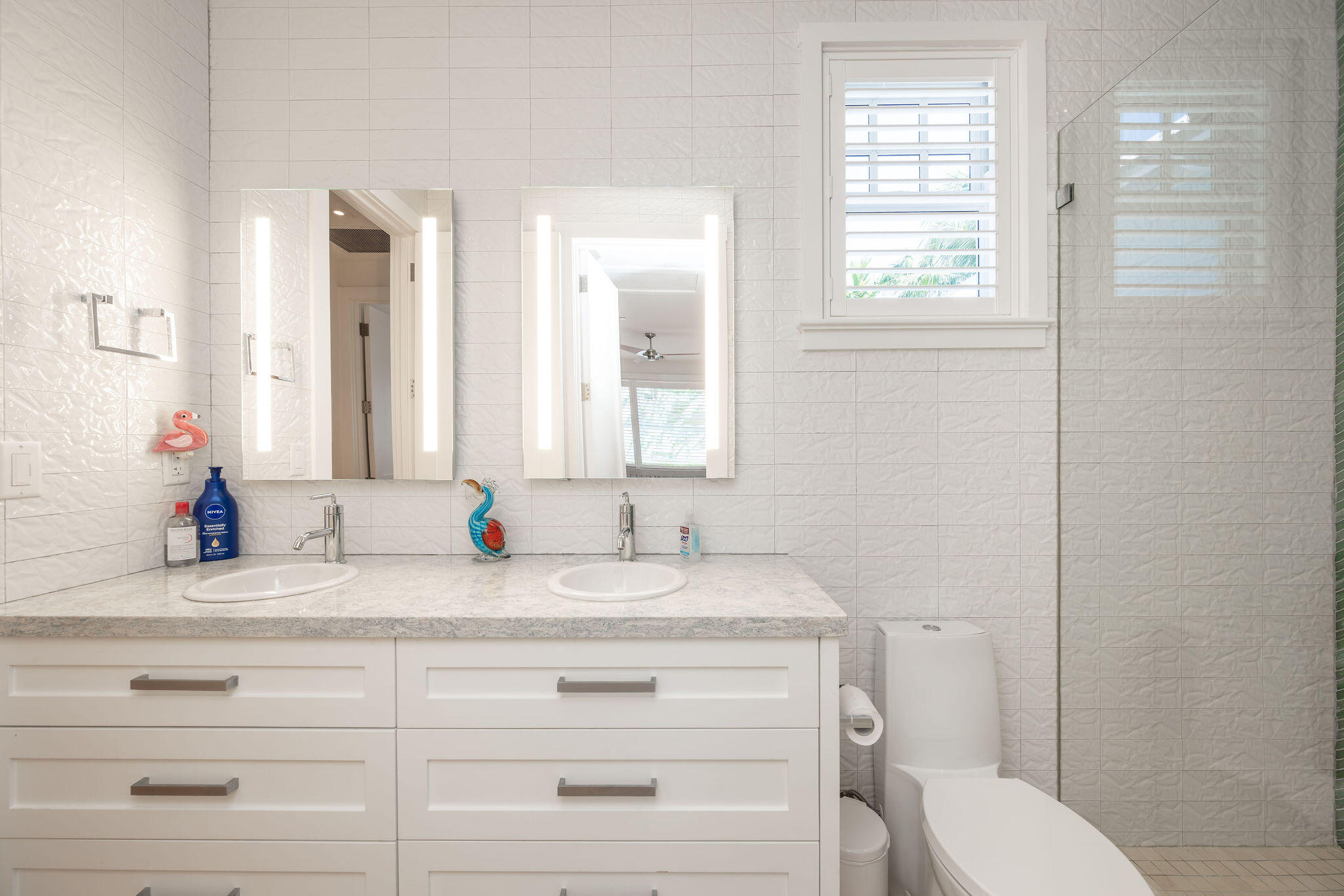 Bathroom with a double-sink vanity, illuminated mirrors, shuttered window, and glass shower.