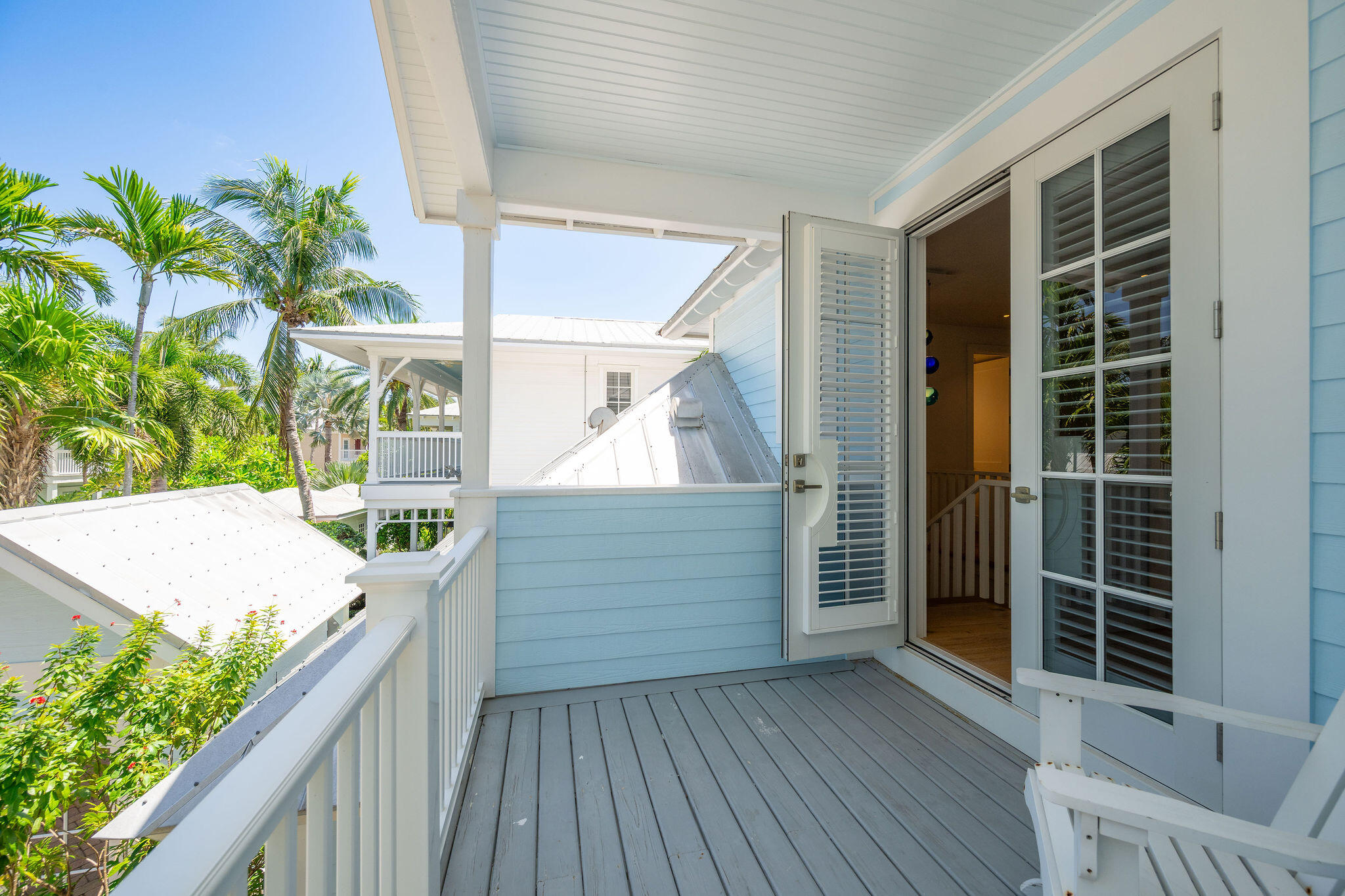 Covered balcony with gray decking and open louvered doors, overlooking palm trees and neighboring homes.