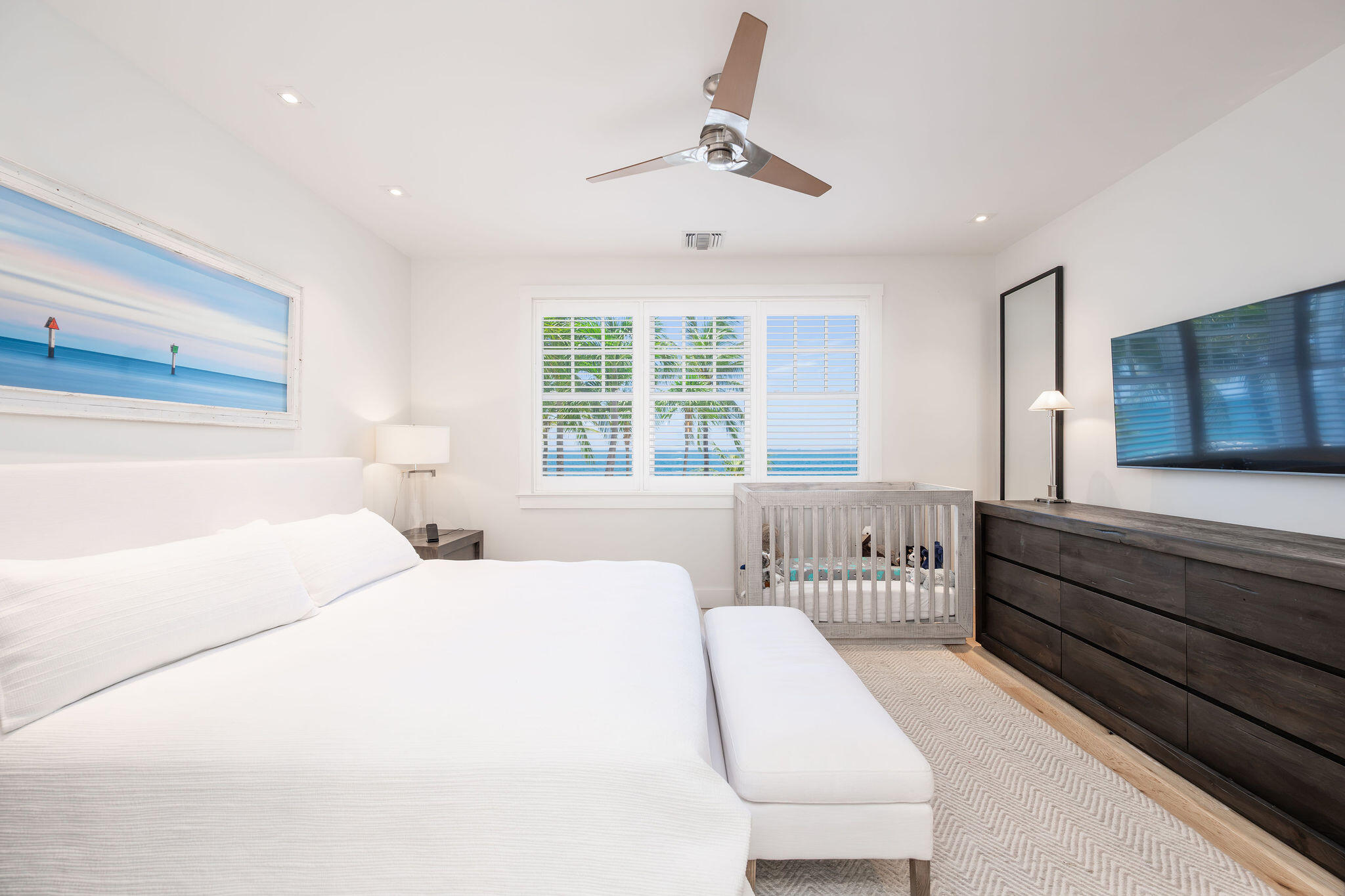 Bedroom with a white bed and gray crib, overlooking palm trees and blue water through shuttered windows.