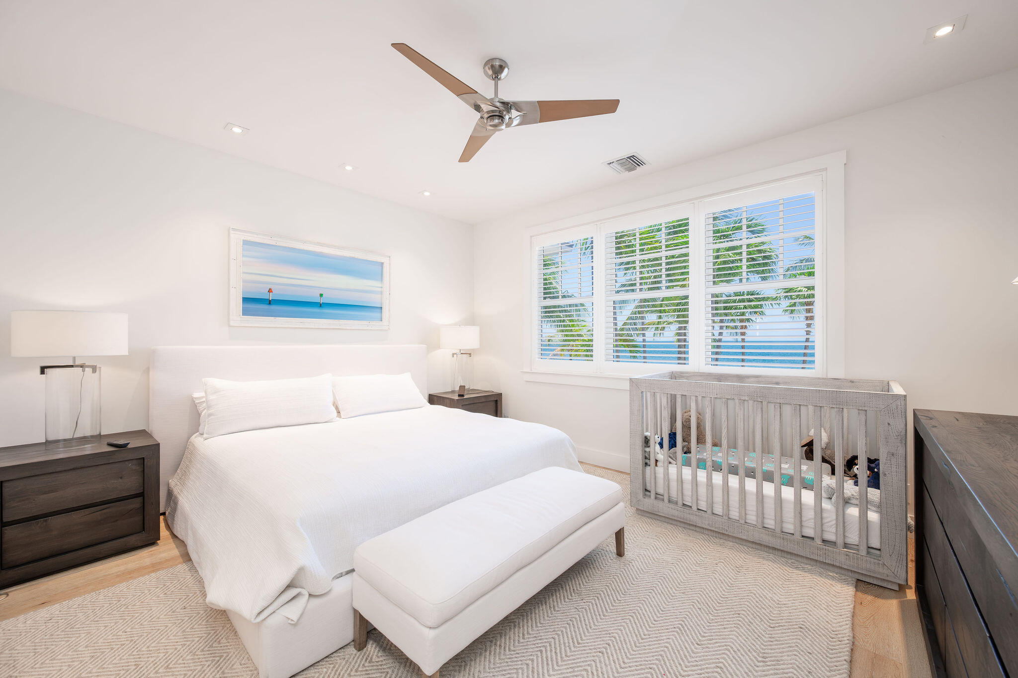 Bright bedroom with a white bed, gray crib, and shuttered windows overlooking palm trees and water.