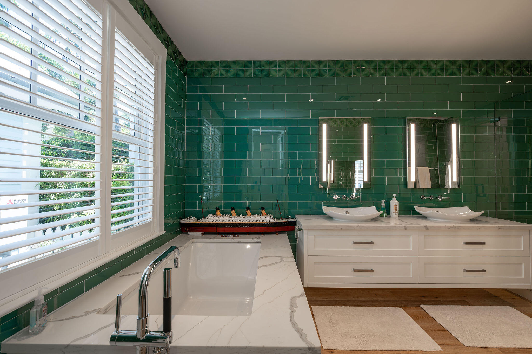 Bathroom with dark green tile, a marble tub surround, double vanity, and plantation shutters.