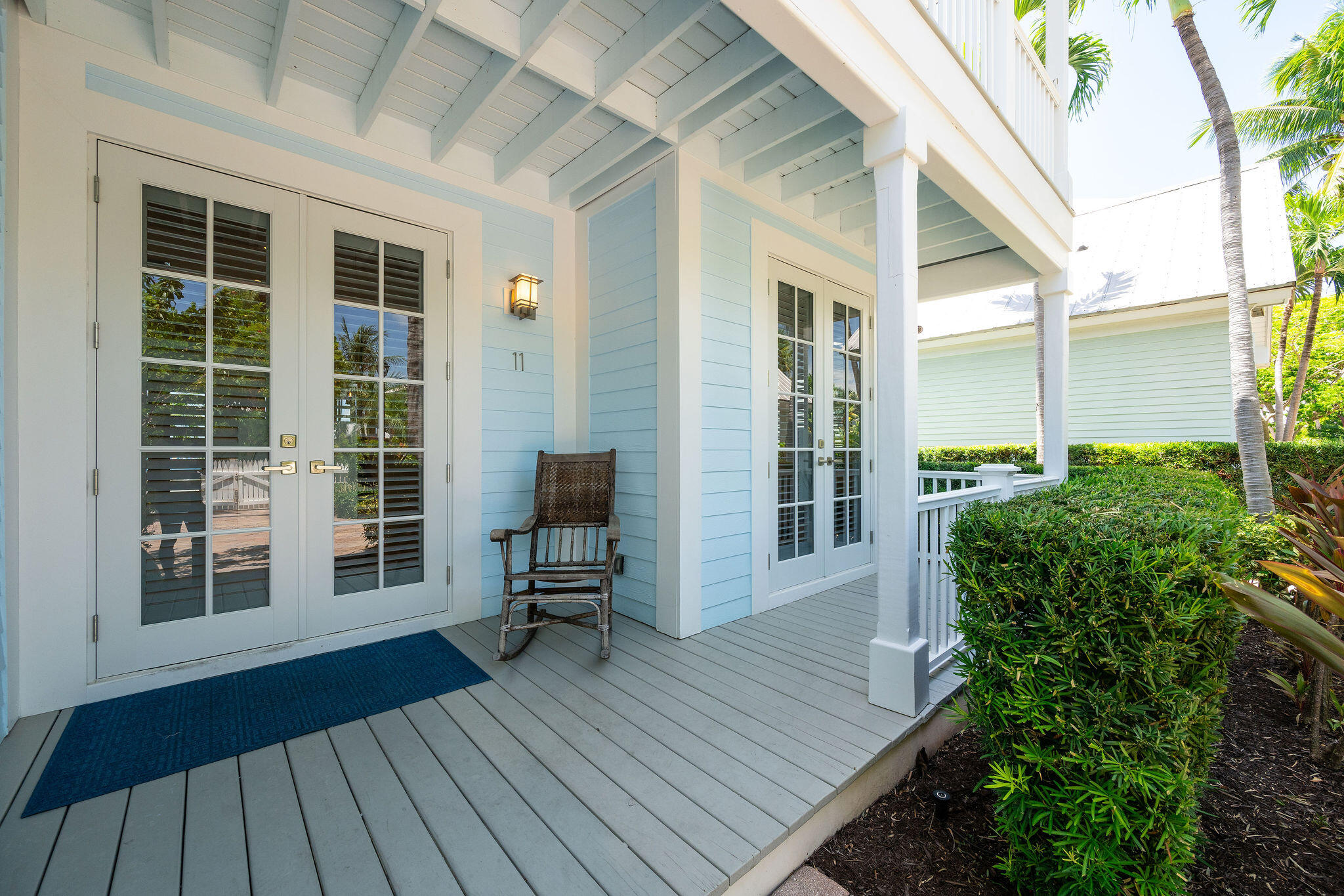 Covered porch with pale blue siding, shuttered French doors, a rocking chair, and tropical landscaping.