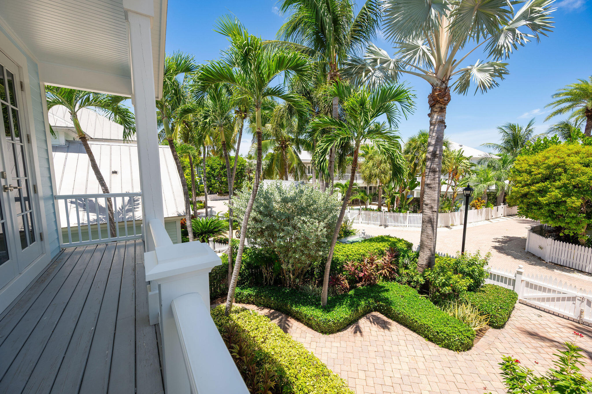 Porch overlooking palm trees, tropical gardens, and a paved path beside white picket fences.