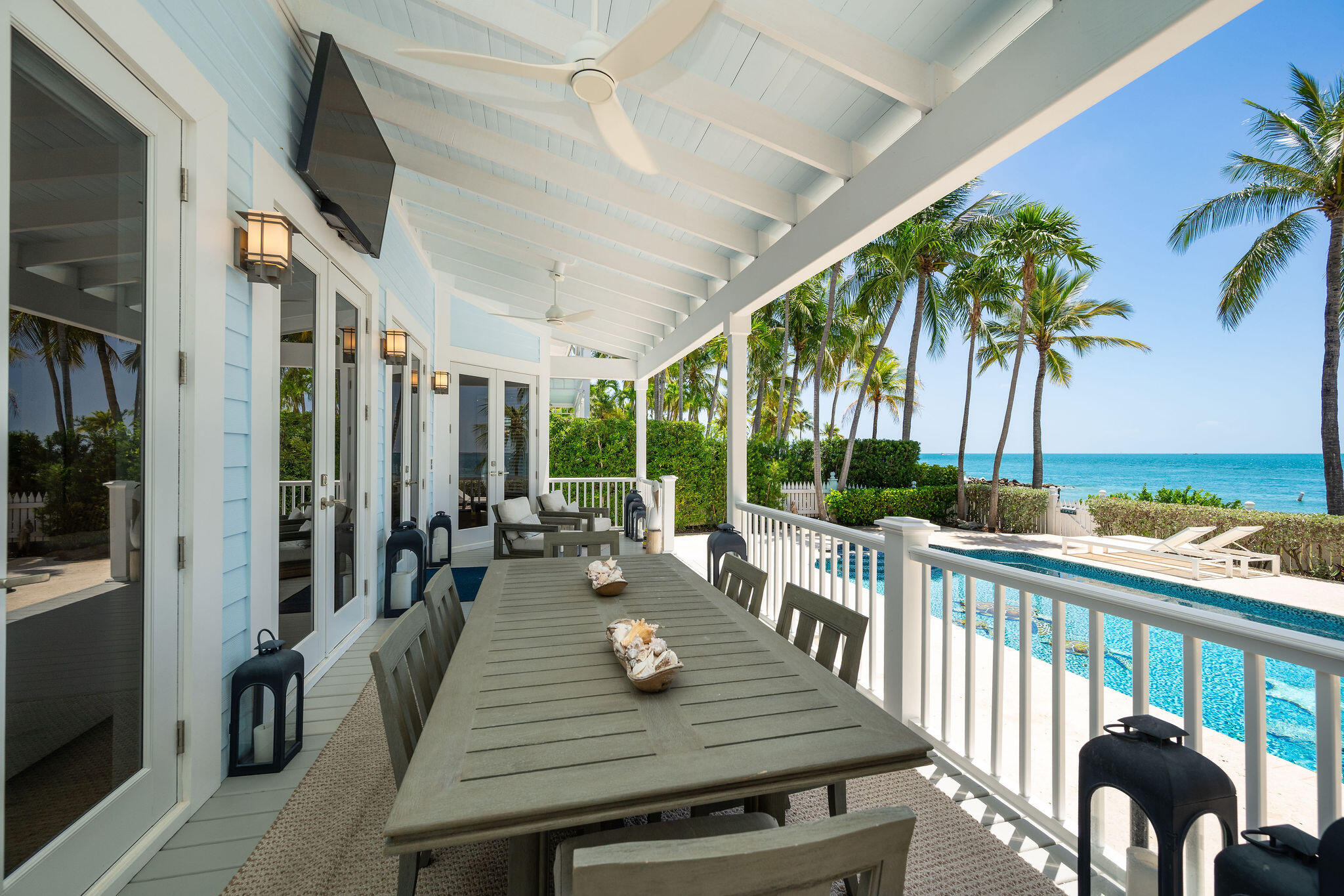 Oceanfront porch with a long dining table overlooking a pool, palm trees, beach, and turquoise water.