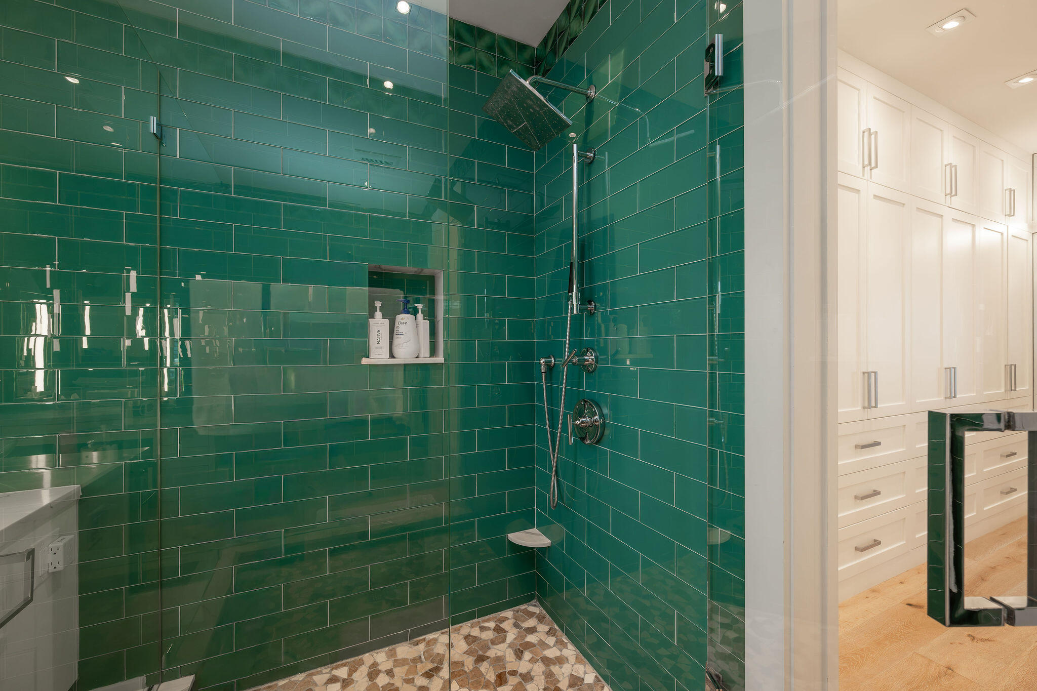 Walk-in shower with glossy green tile, rainfall showerhead, and pebble-mosaic floor beside white bathroom cabinetry.
