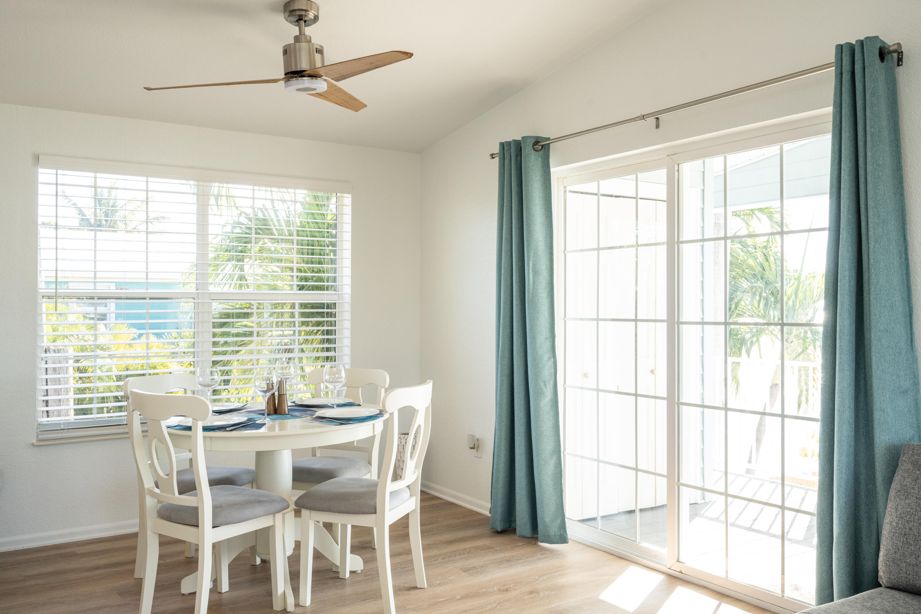 Dining room with a set white table, teal curtains, and sliding glass doors overlooking palm foliage.