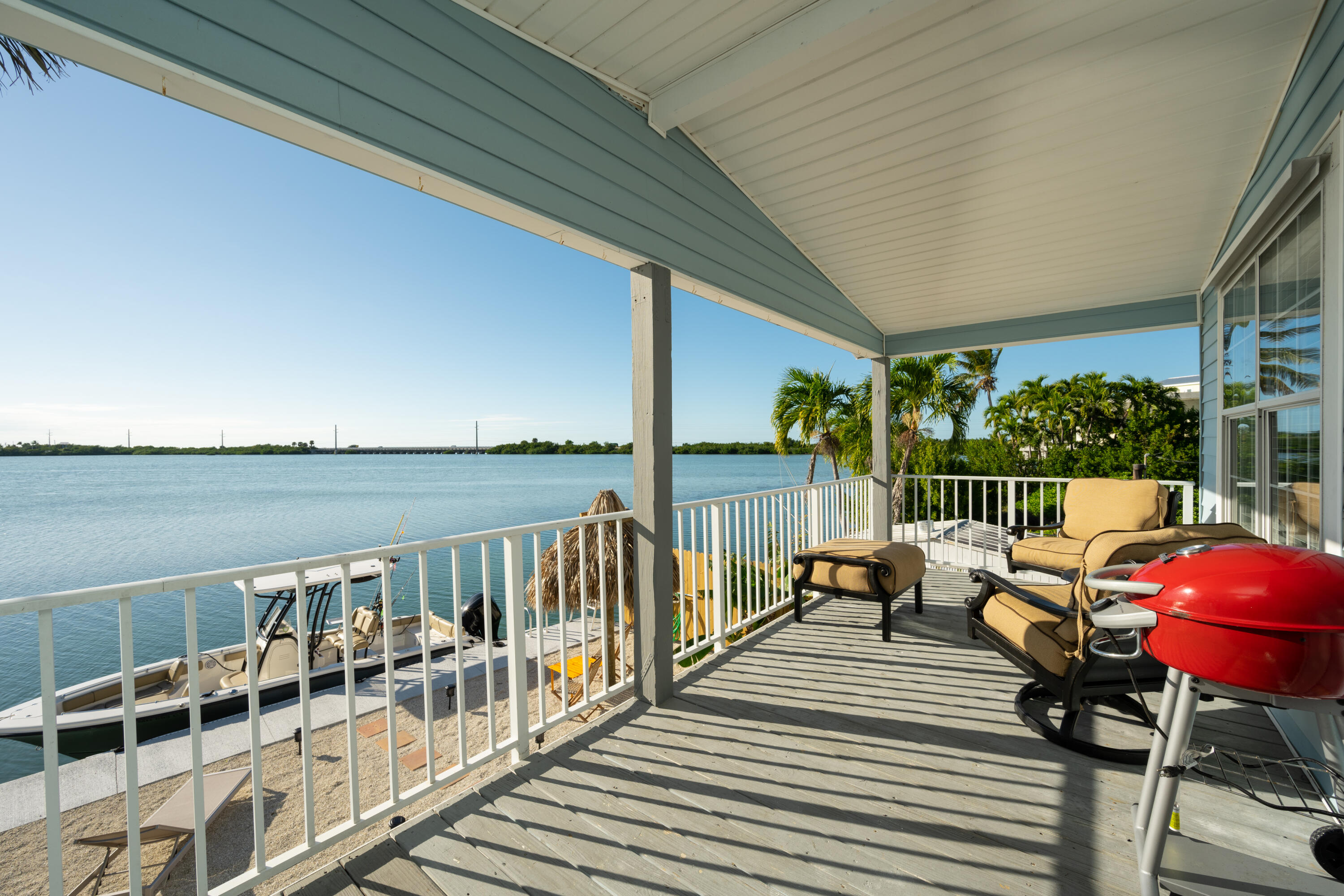 Covered waterfront deck with lounge chairs and a red grill overlooking calm water and a docked boat.