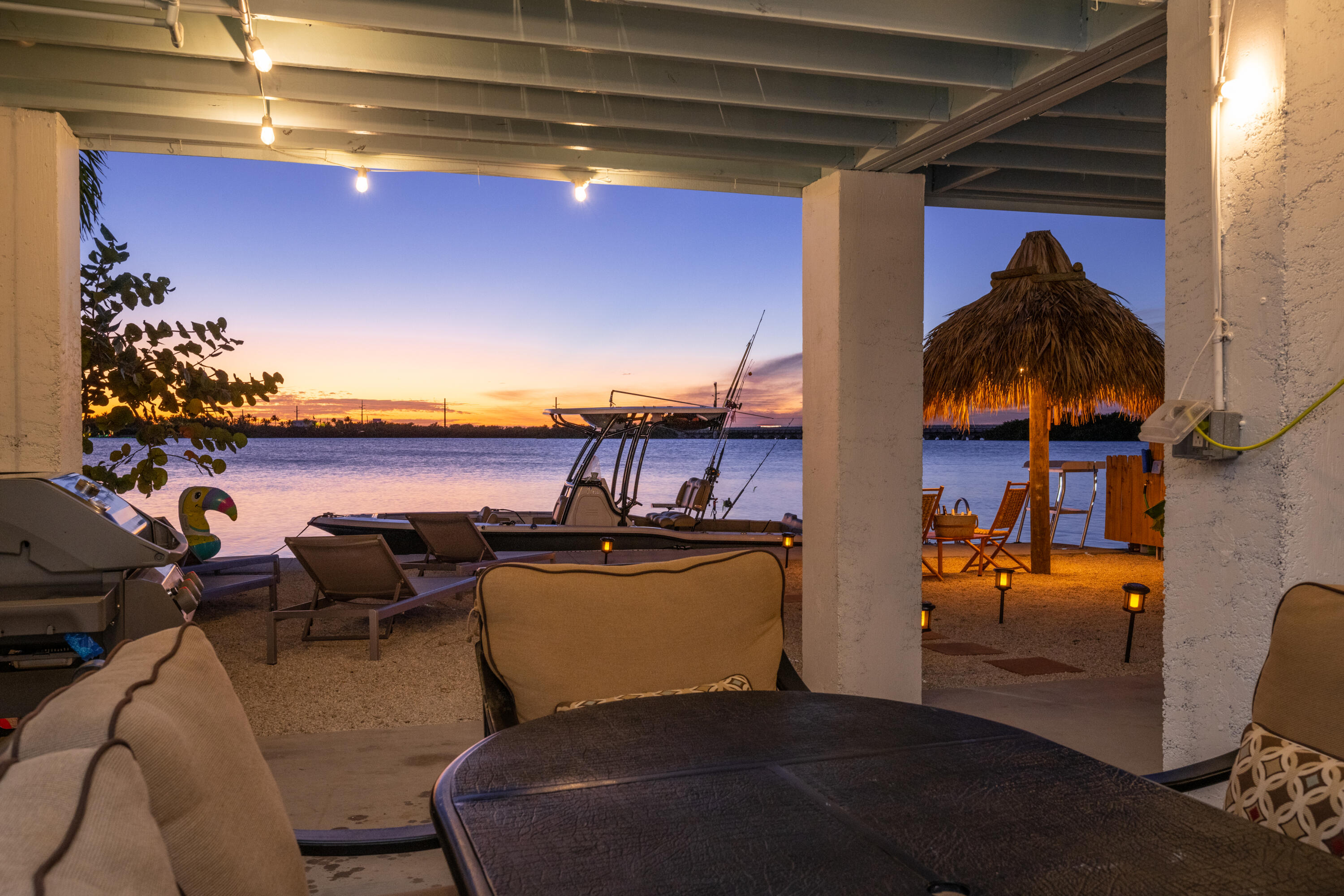Covered waterfront patio with a thatched tiki hut, docked boat, and sunset view.