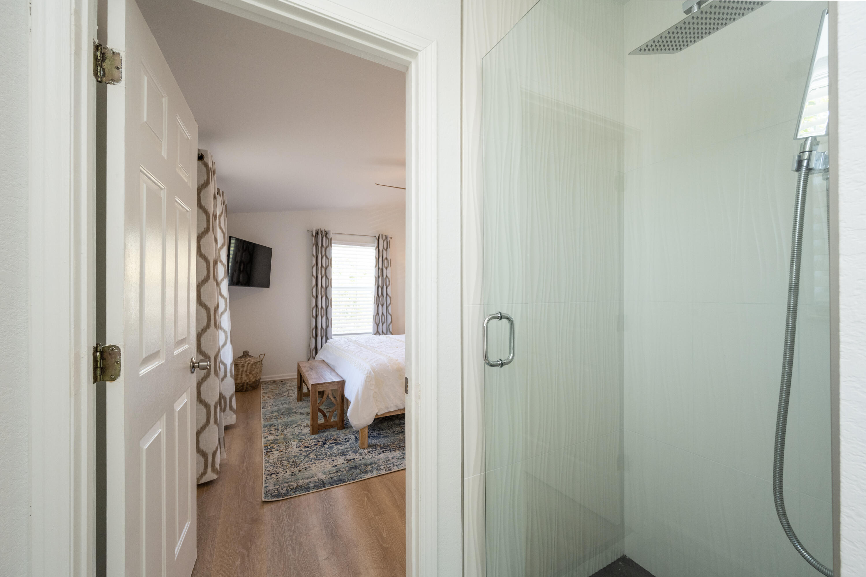 Walk-in shower beside a furnished bedroom with a white bed and patterned rug.