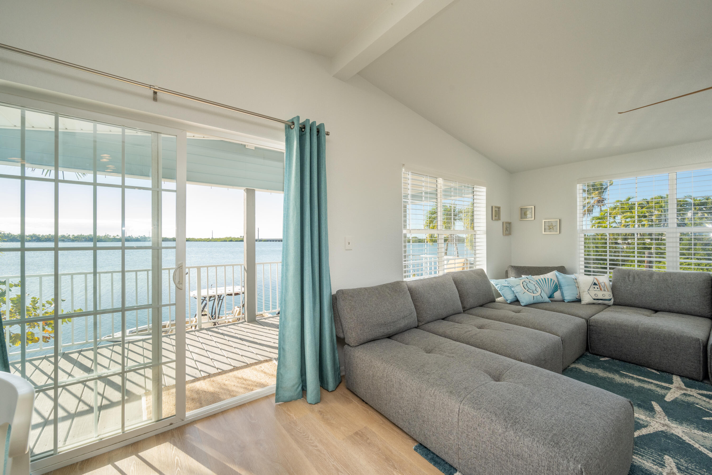Waterfront living room with a gray sectional, turquoise curtains, and sliding doors opening to a balcony.