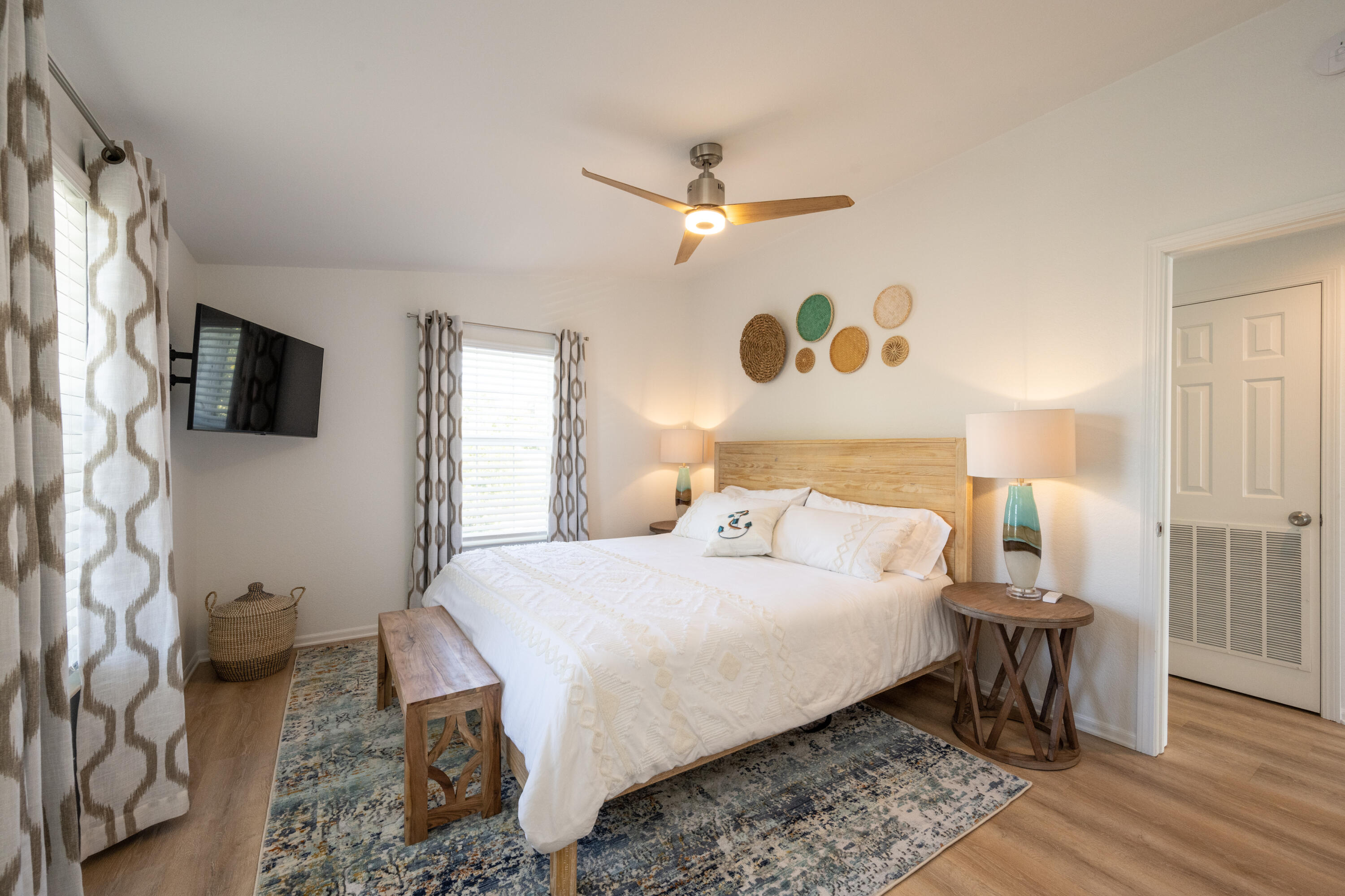 Bedroom with a white bed, light wood headboard, patterned rug, and woven wall decor.