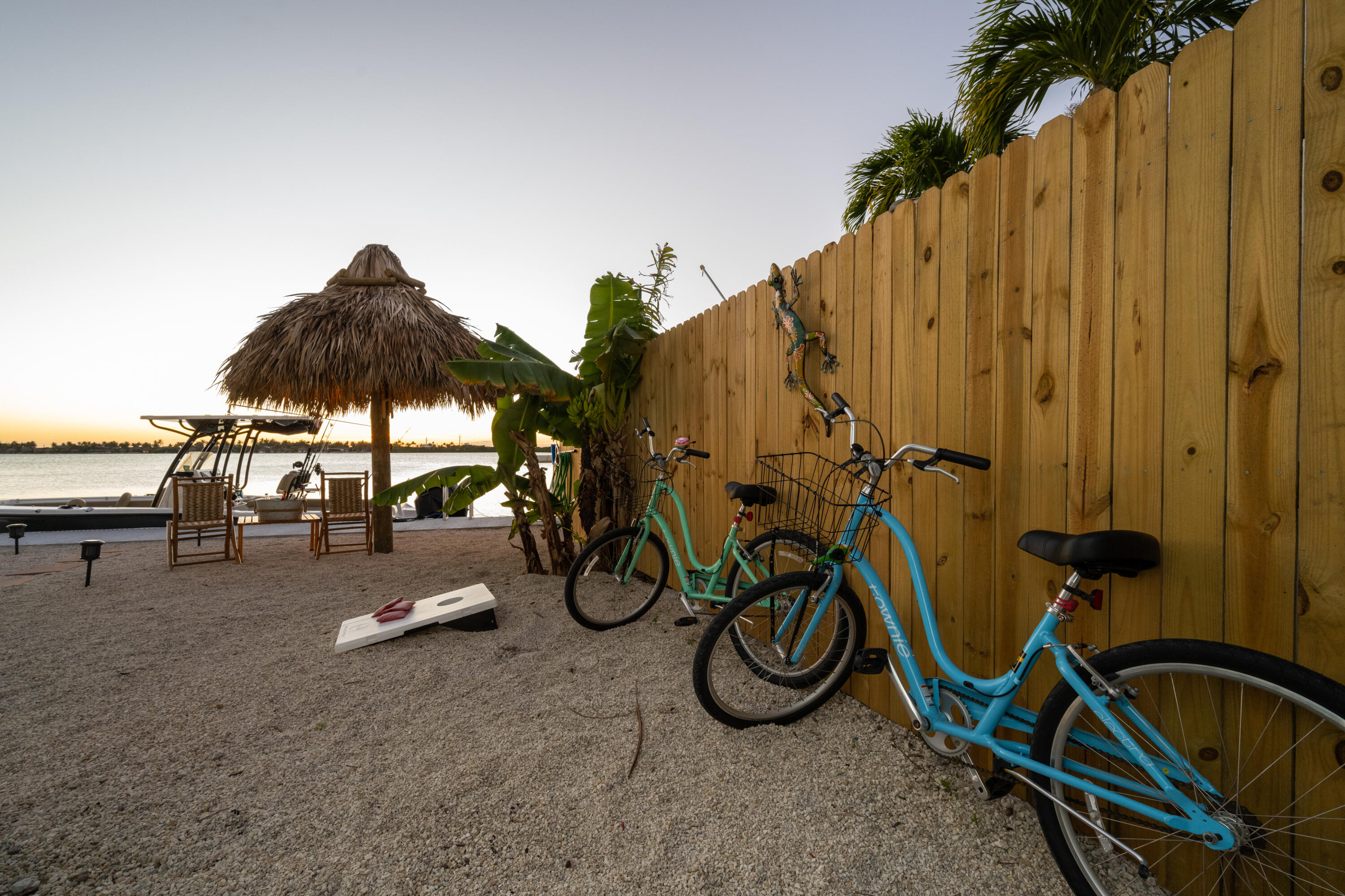 Waterfront patio with turquoise bicycles, a thatched tiki umbrella, and chairs beside the bay at sunset.