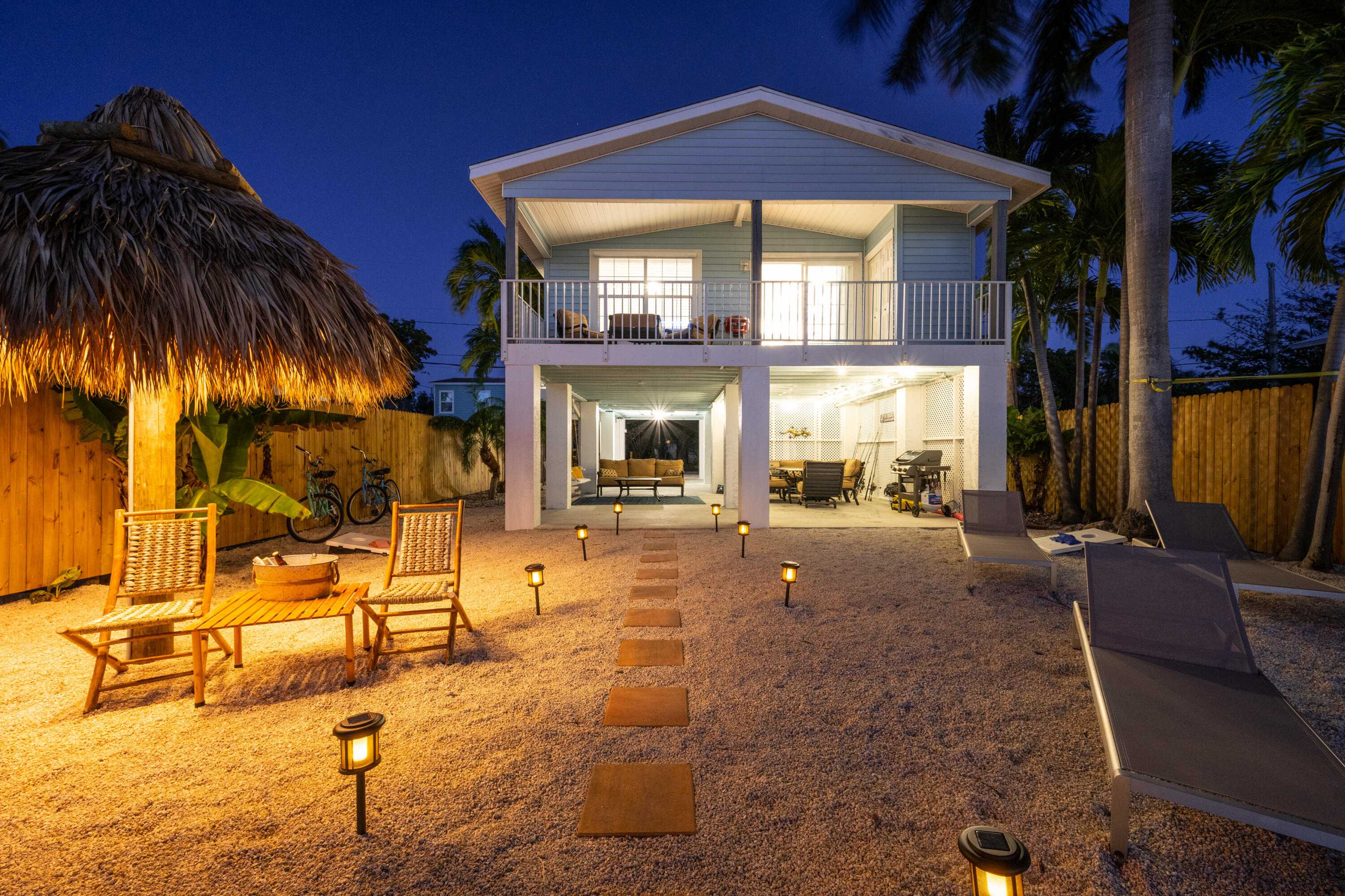 Raised coastal home at night with a covered balcony, tiki shelter, and illuminated gravel yard.