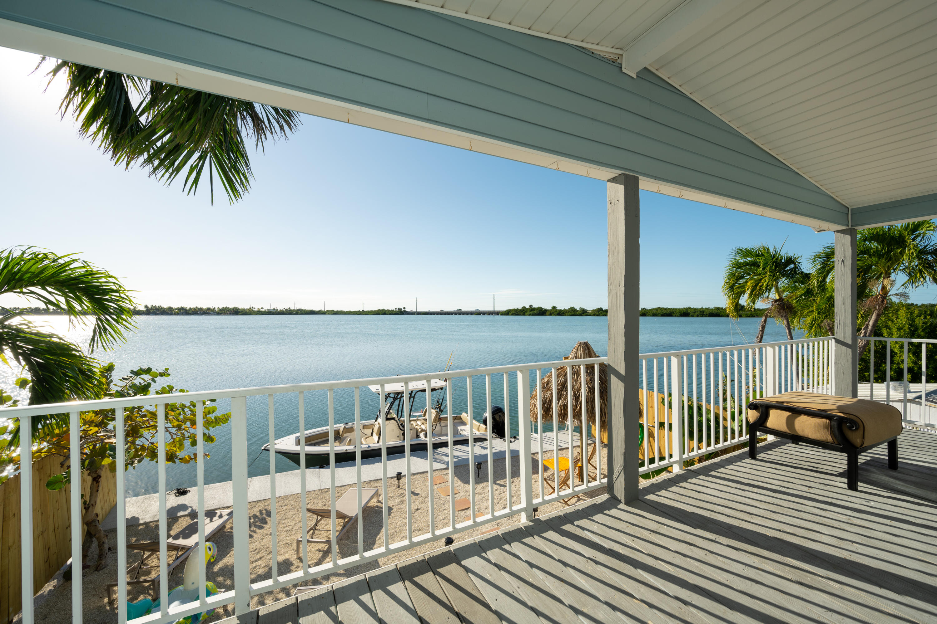 Covered deck overlooking calm water, with a boat below and palm trees along the shore.