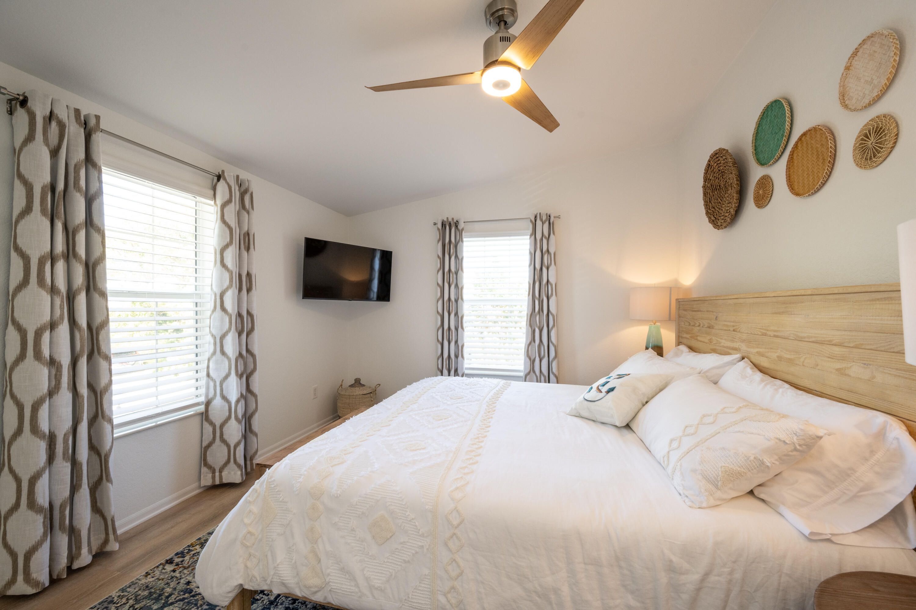 Bedroom with a white bed, natural wood headboard, curtained windows, and woven wall decor.