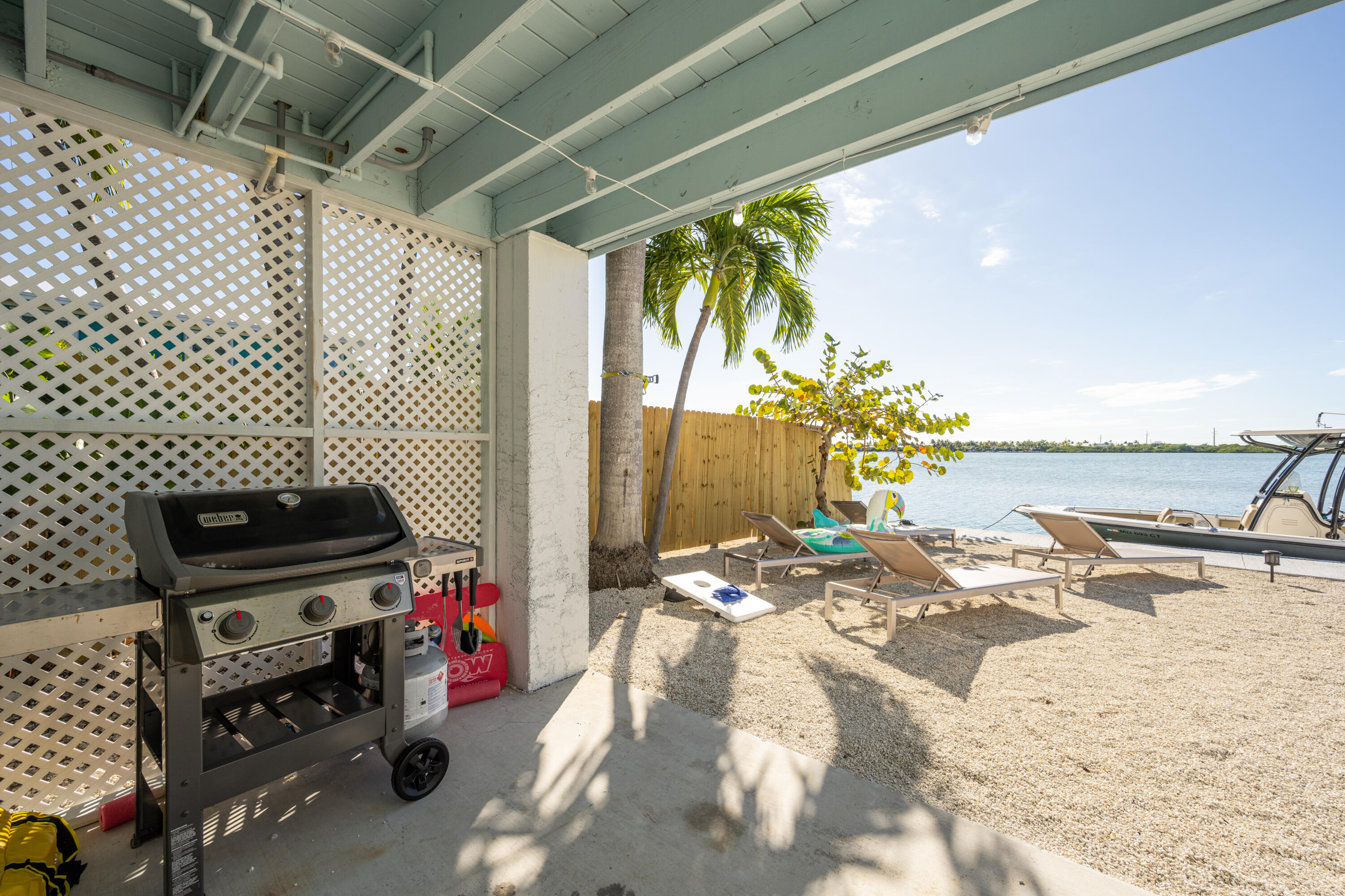 Covered patio with a Weber grill overlooking sandy waterfront seating and a boat.