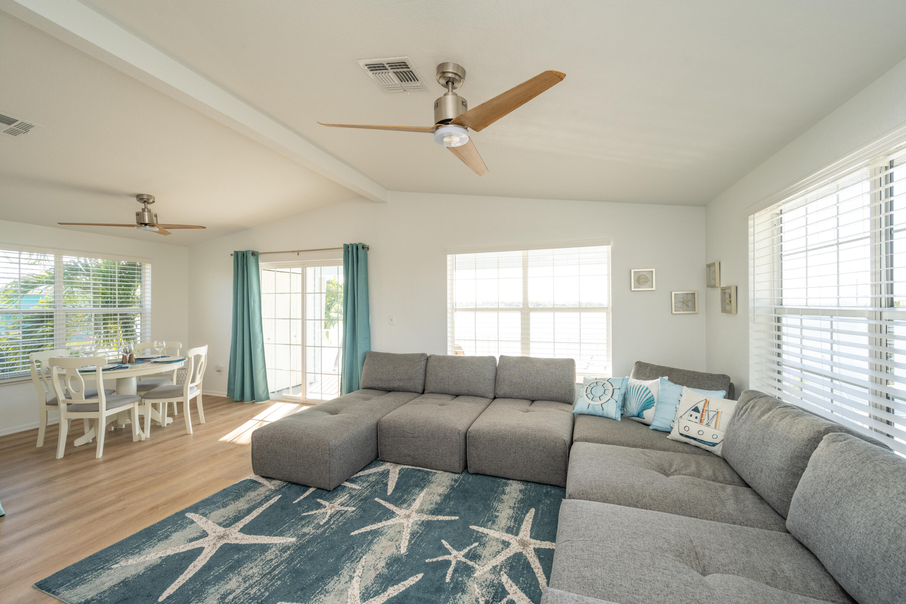 Open-plan living and dining room with a gray sectional, blue starfish rug, and water views.