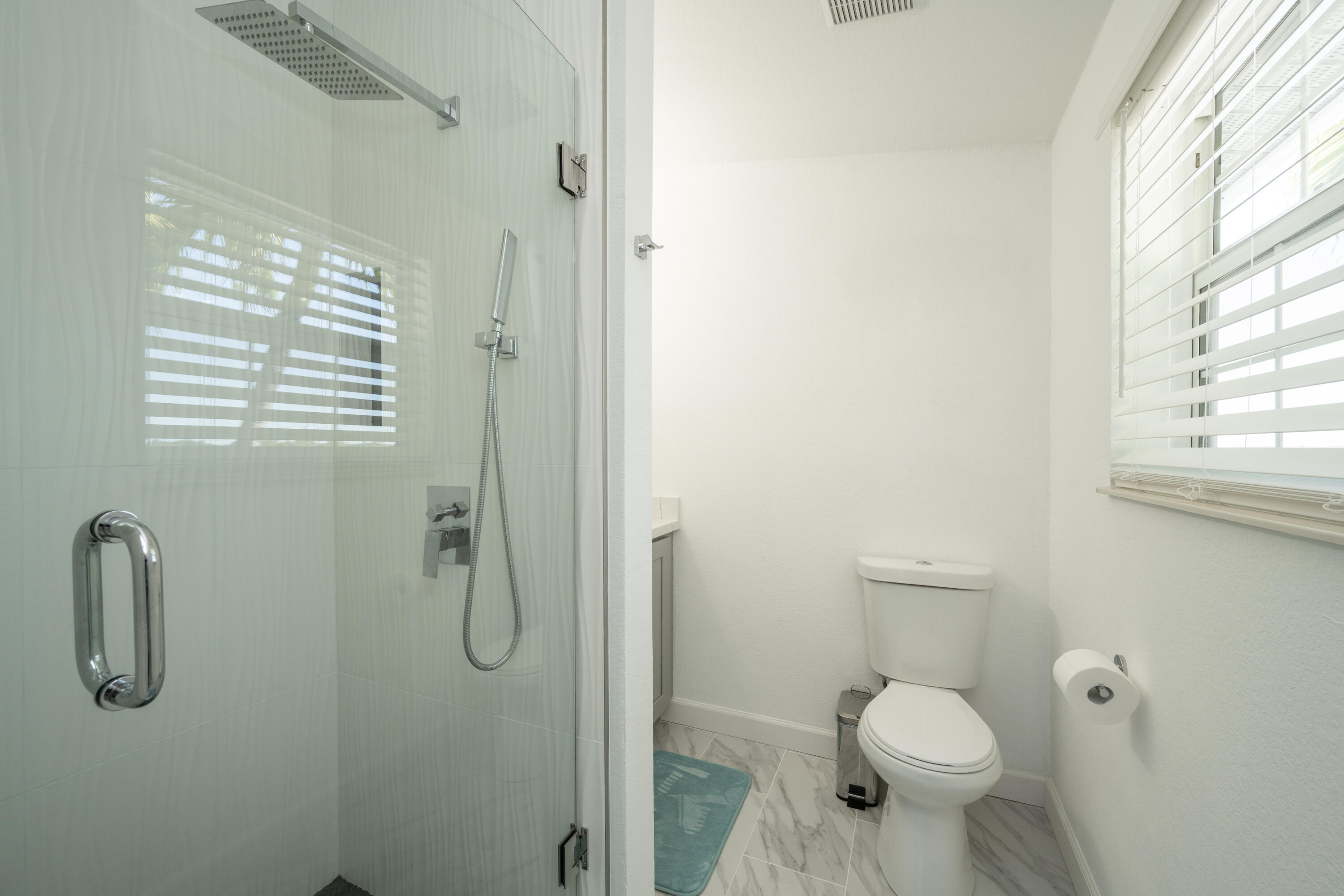 Bathroom with a glass shower, chrome rainfall showerhead, toilet, and white plantation blinds.