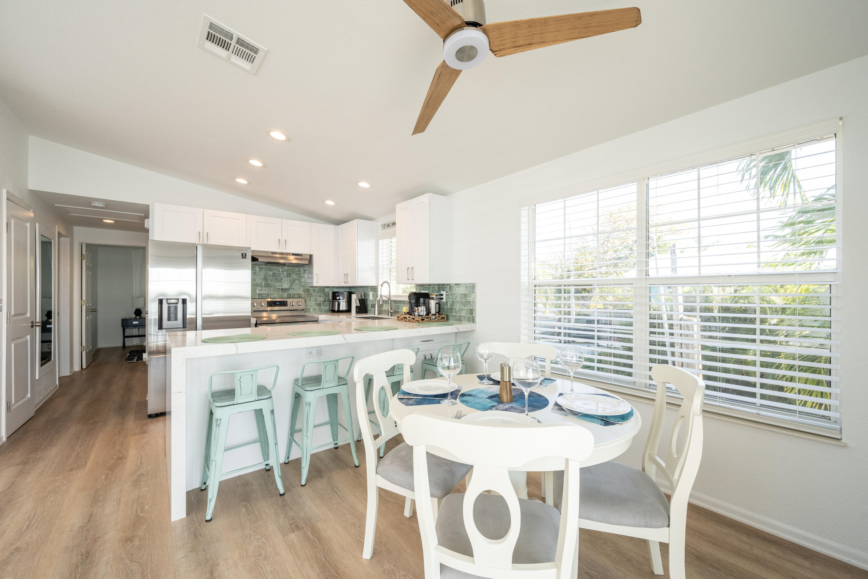 Kitchen and dining area with green tile backsplash, mint stools, and a dining table by a palm-framed window.
