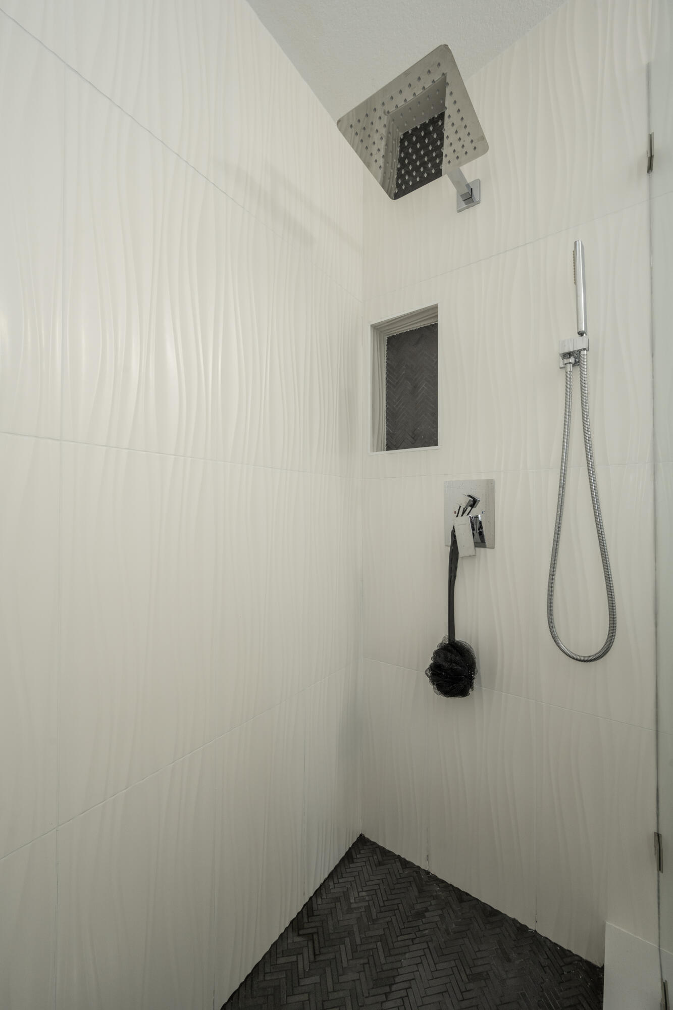 Modern shower with a square rainfall head, handheld wand, recessed niche, and dark herringbone floor.
