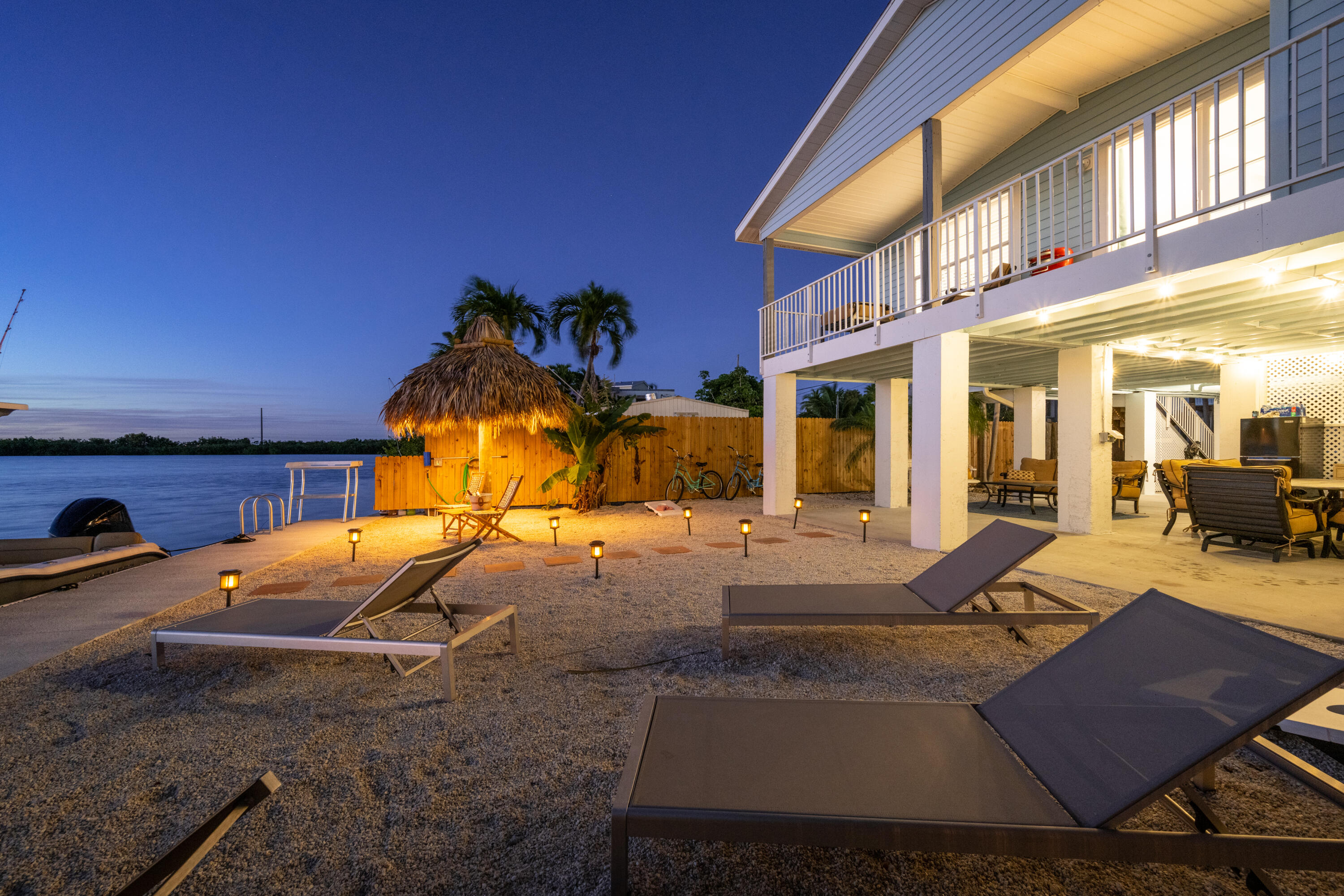 Waterfront home at dusk with a lit tiki hut, gravel lounge area, and covered patio beside calm water.