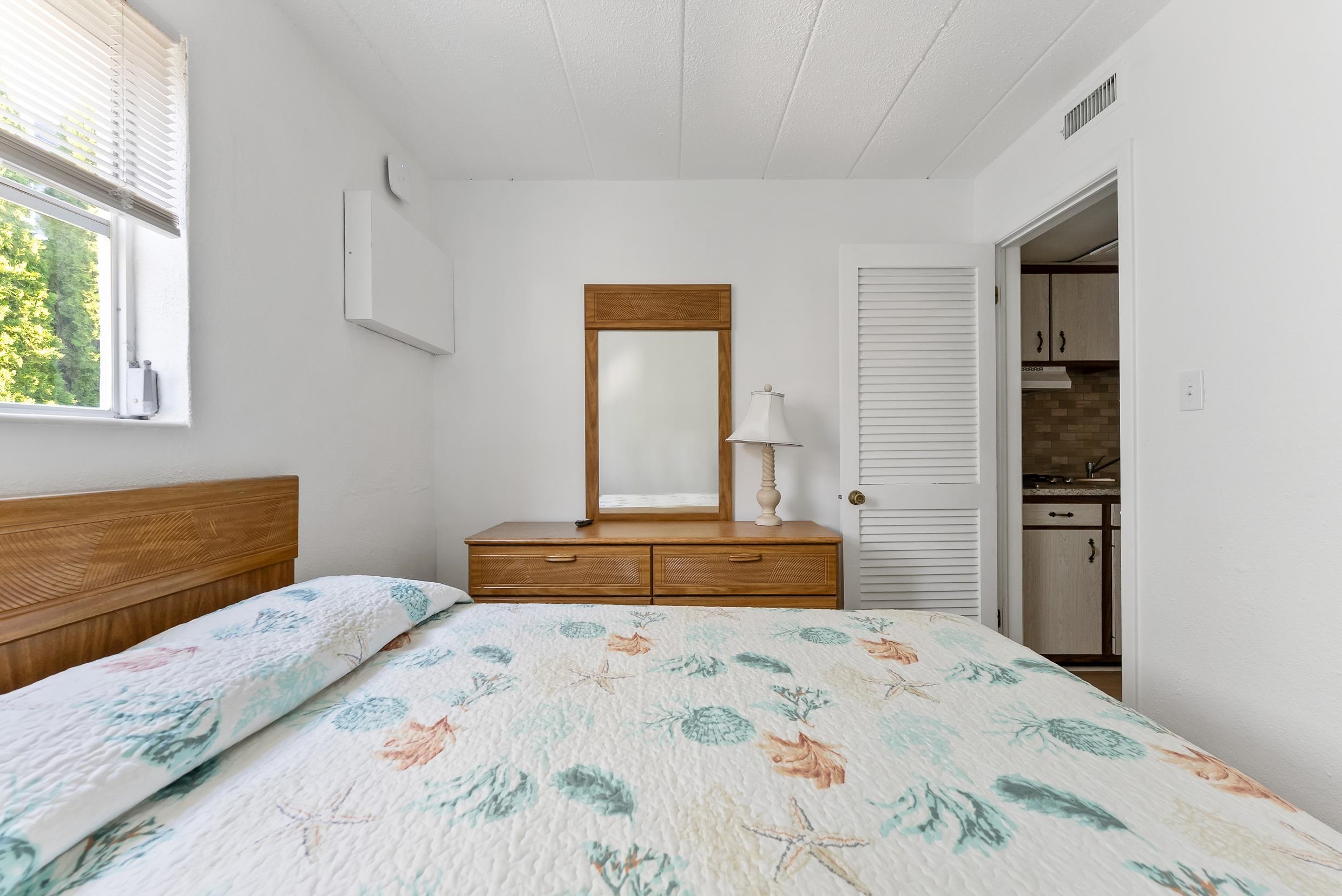 Bedroom with a seashell-patterned quilt, wooden dresser and mirror, and window beside the bed.