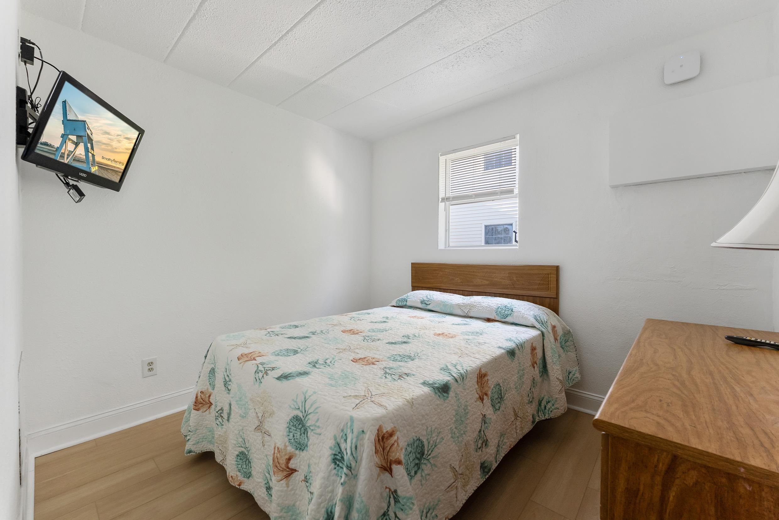 Furnished bedroom with a sea-life patterned quilt, wall-mounted TV, and wooden dresser.