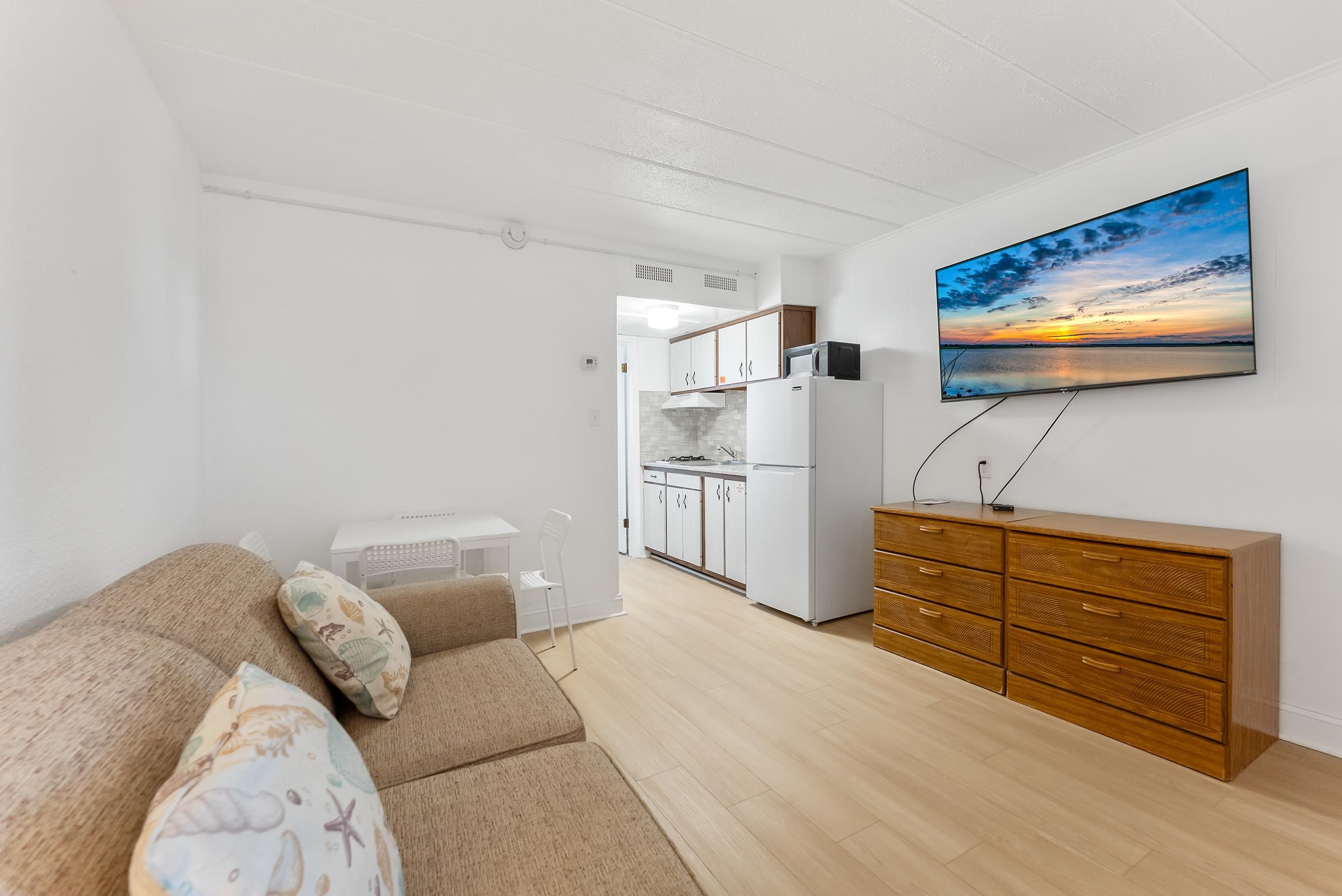 Furnished living area with beige sofa, compact kitchenette, and wall-mounted television above a wooden dresser.