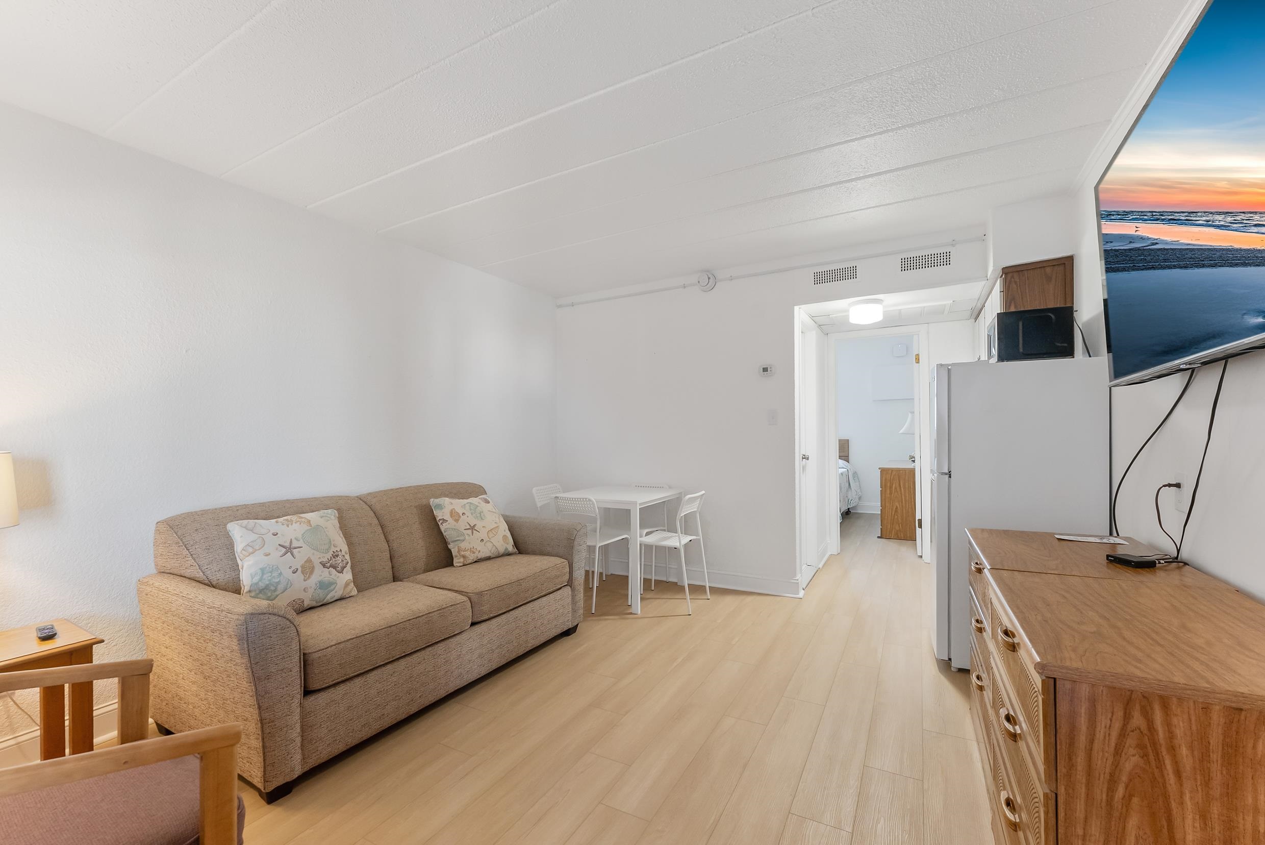 Furnished condo living room with tan sofa, small dining table, and TV displaying a beach sunset.