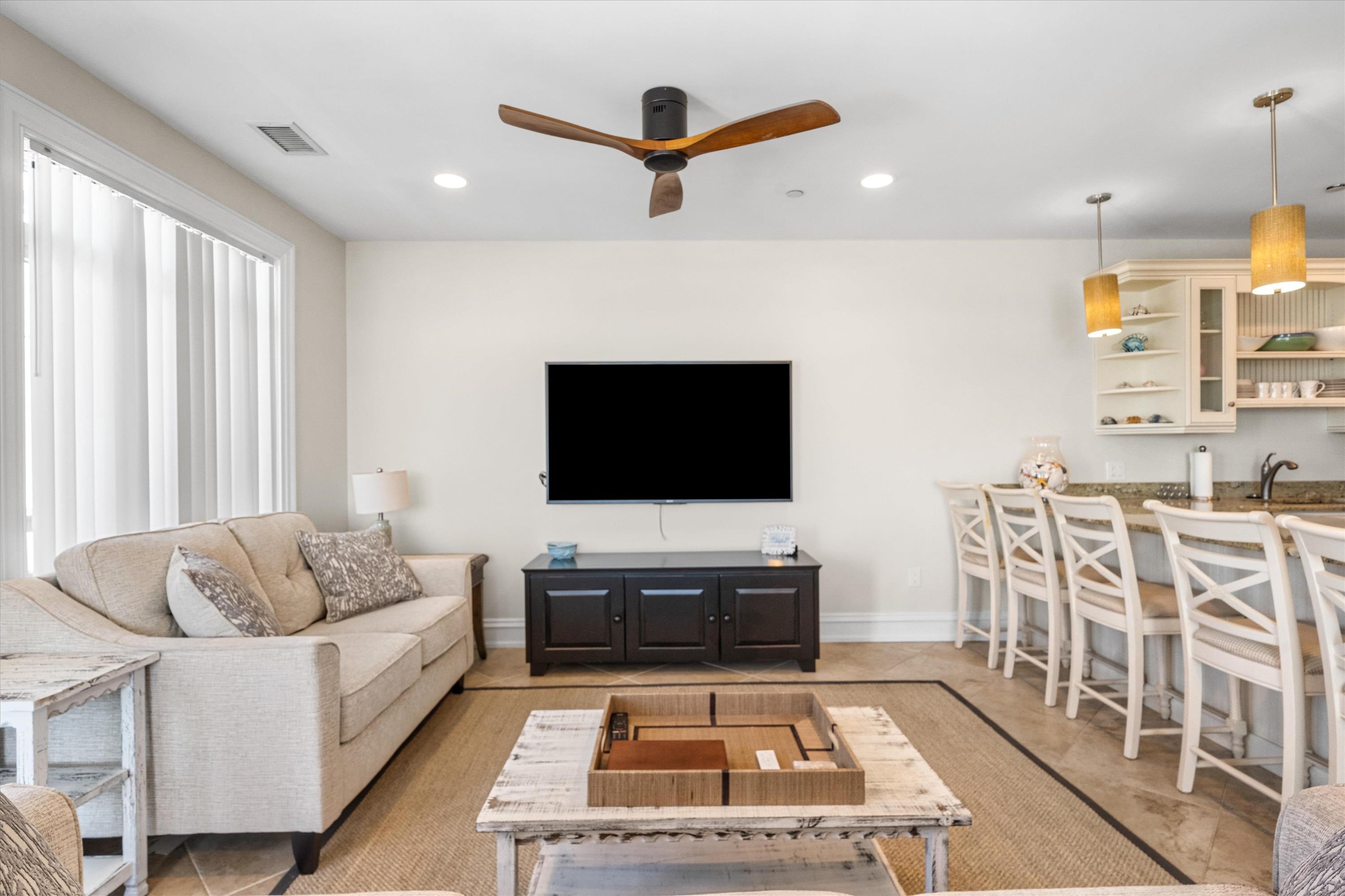 Open-plan living room with beige sofa, wall-mounted TV, and kitchen breakfast bar with white stools.
