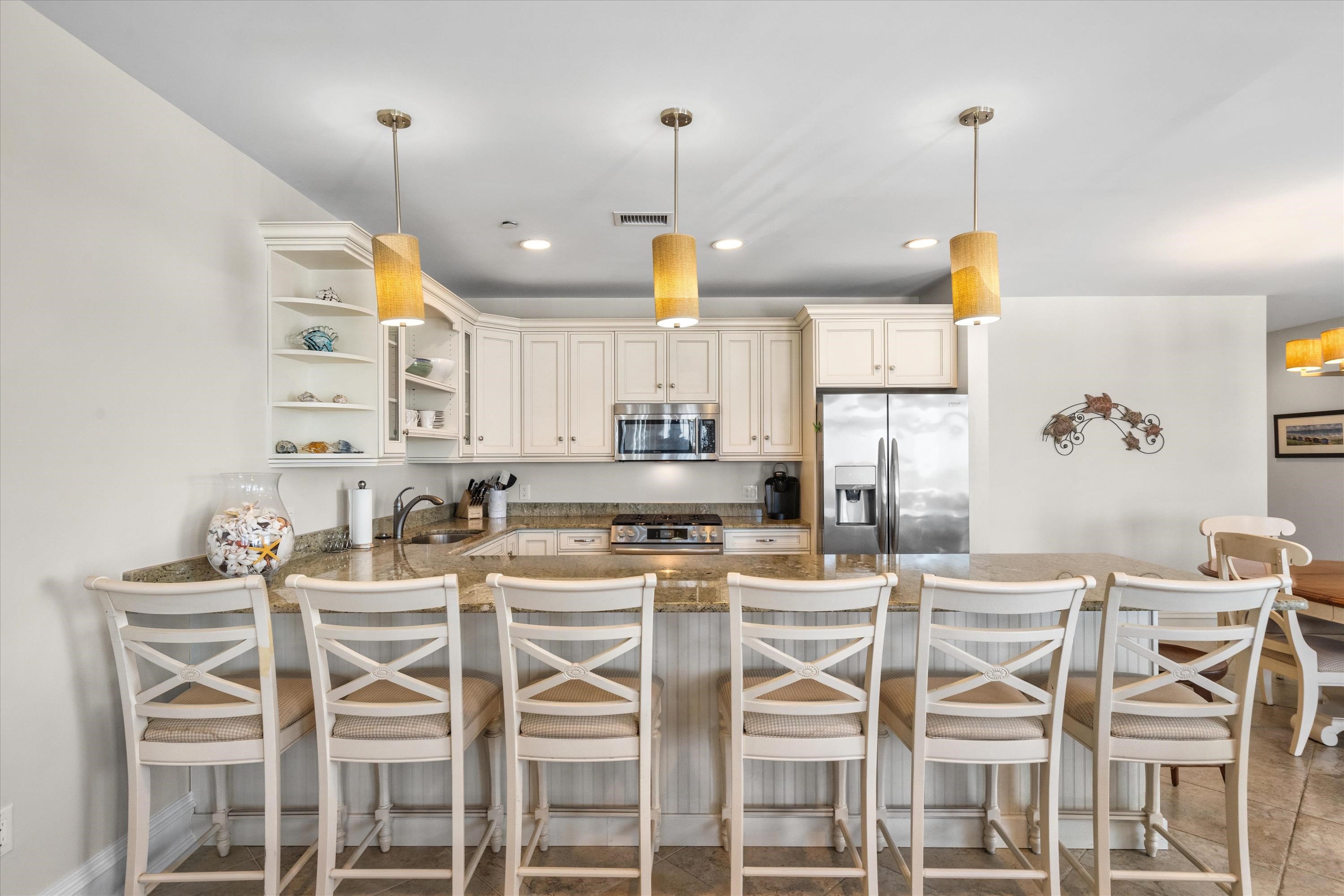 Kitchen with a granite breakfast bar, six white chairs, cream cabinets, and stainless steel appliances.