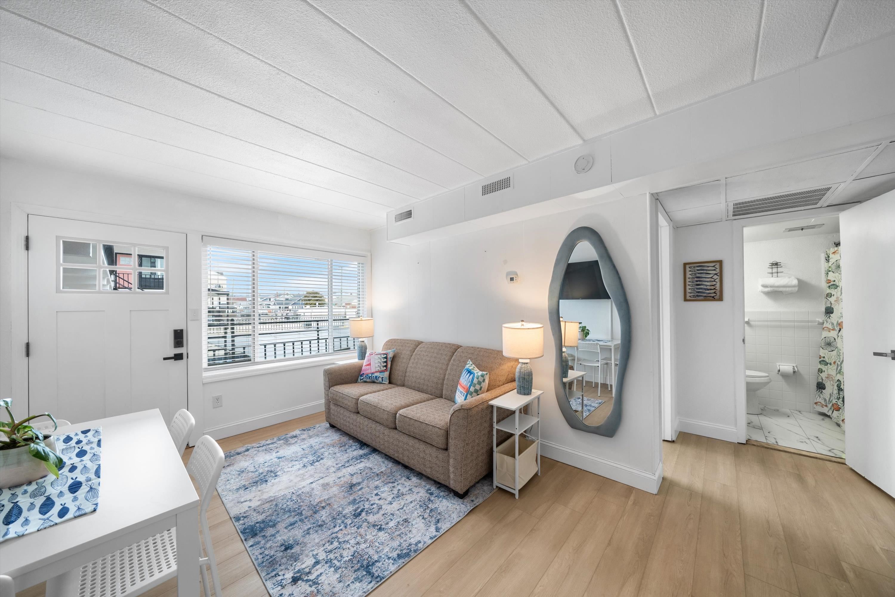Living room with beige sofa, blue patterned rug, and a large window overlooking nearby buildings.
