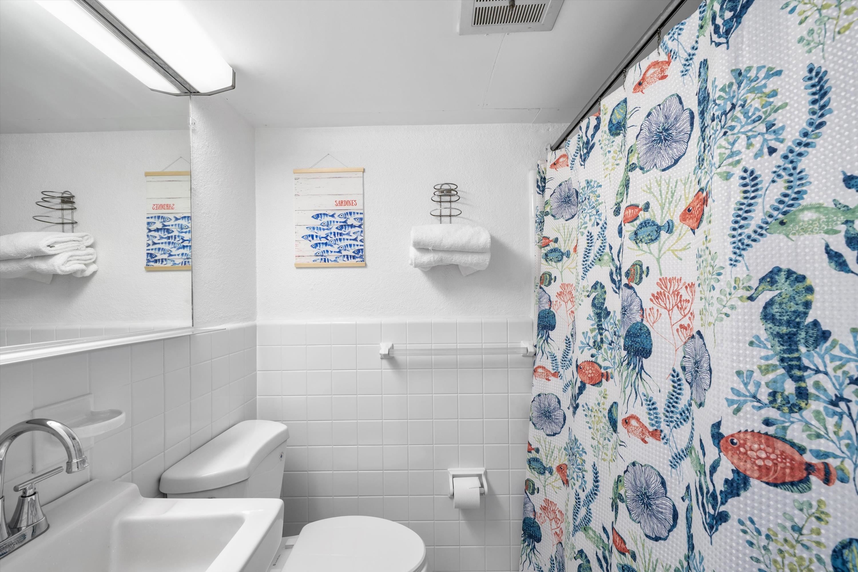 White-tiled bathroom with a colorful sea-life shower curtain and sardine-themed wall art.