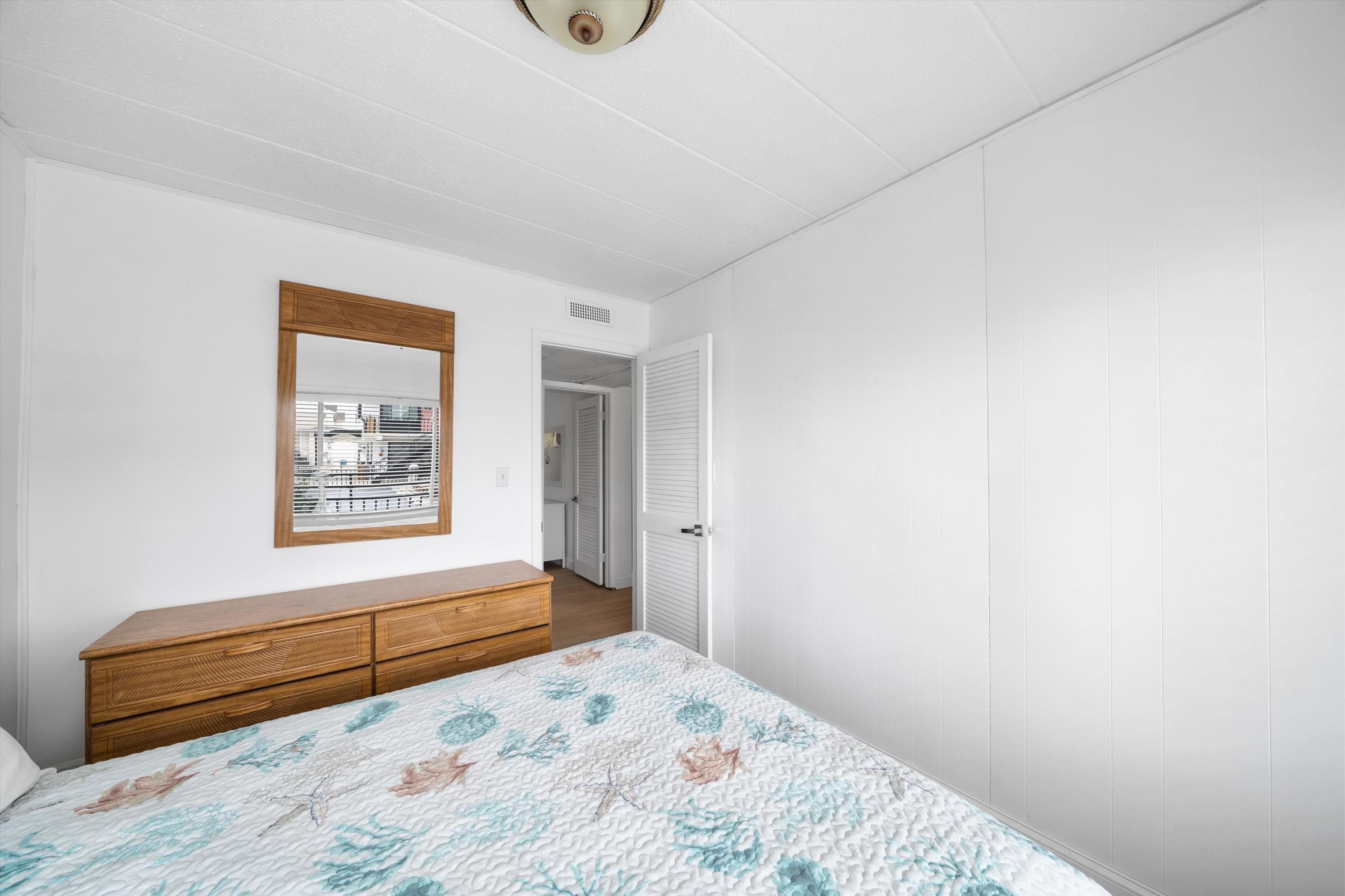 Bedroom with a floral quilt, wooden dresser and mirror, and an open louvered door.