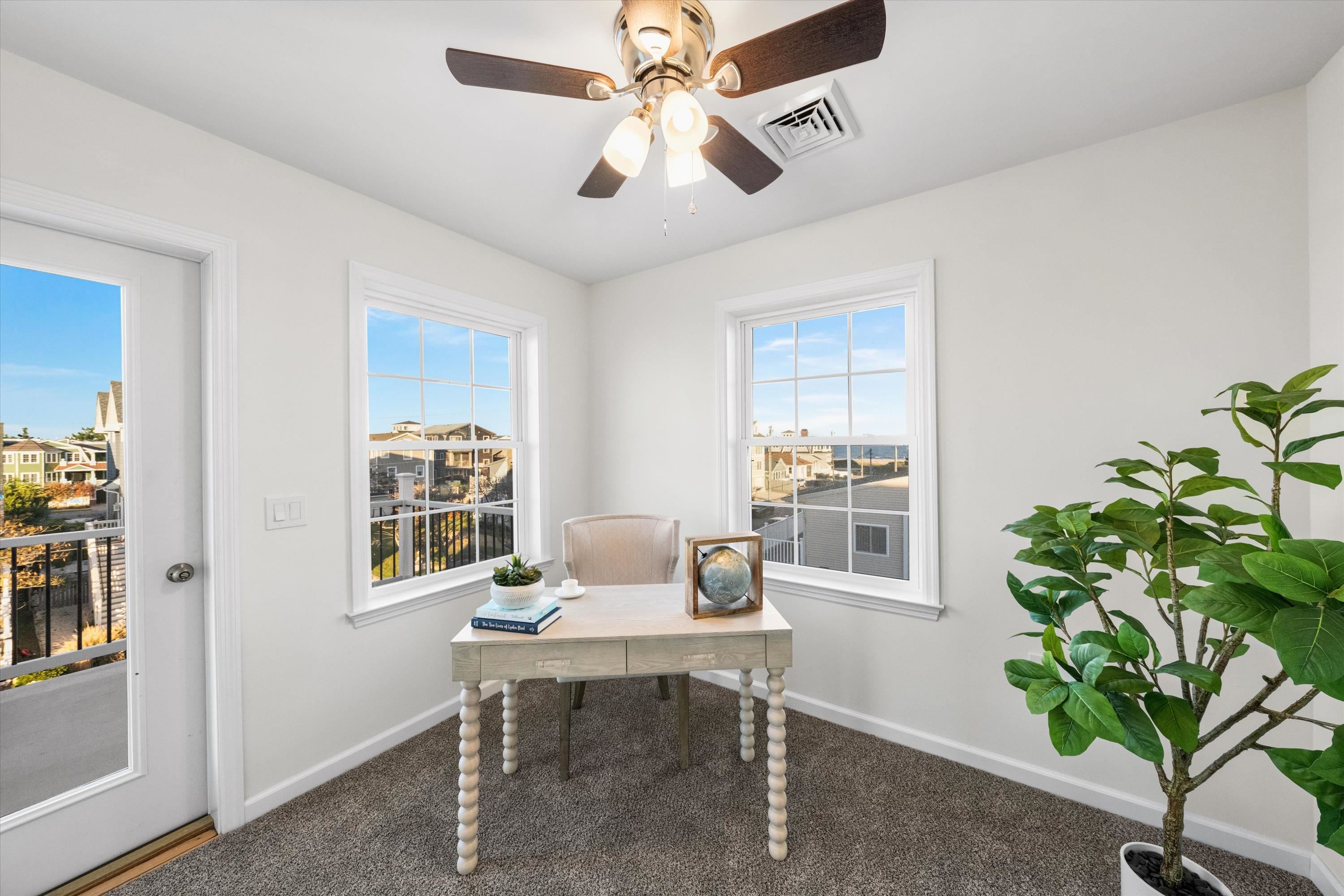 Staged office with a desk and potted plant, overlooking coastal homes and a glimpse of water.
