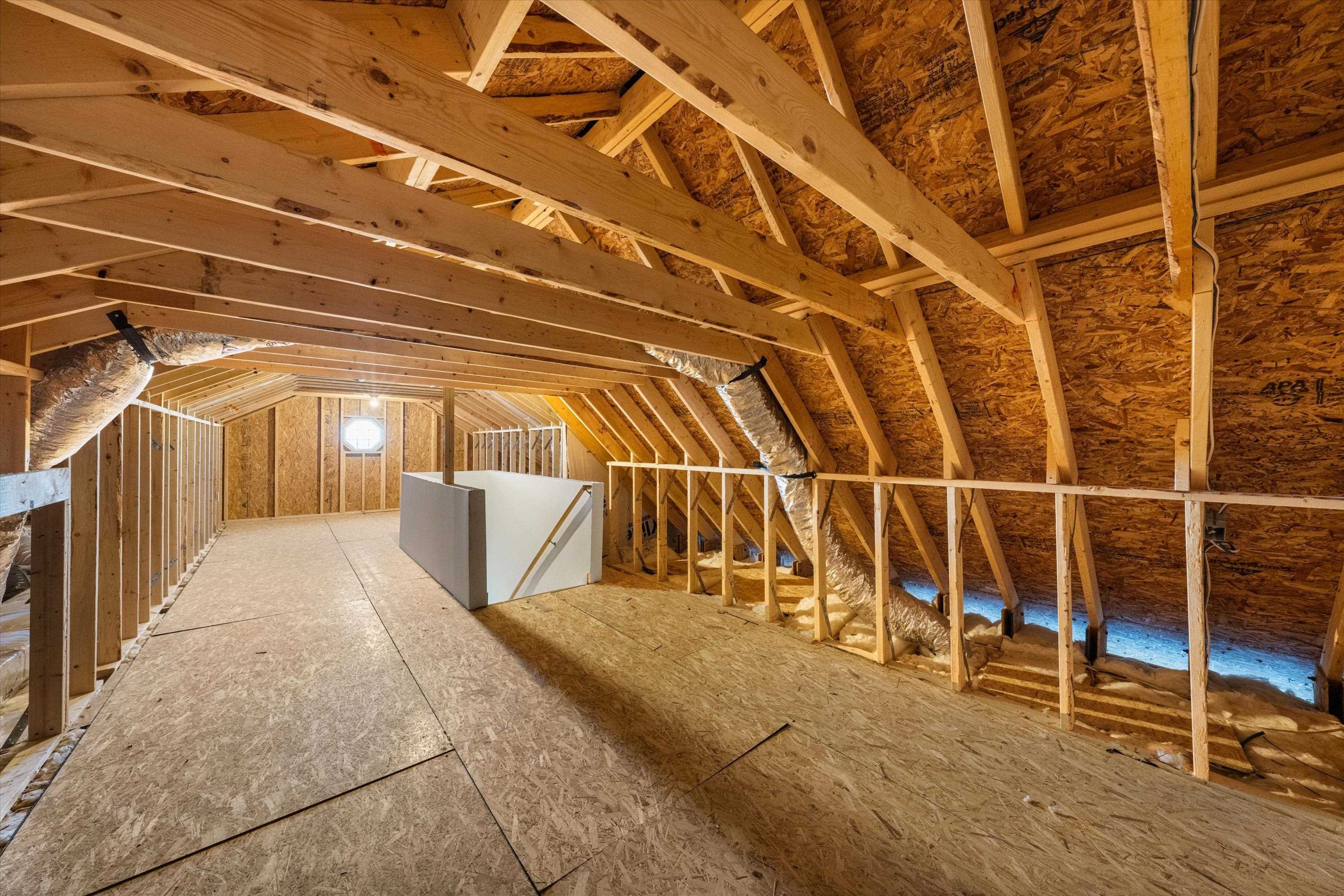 Unfinished attic with exposed roof framing, plywood flooring, ductwork, and a small octagonal window.
