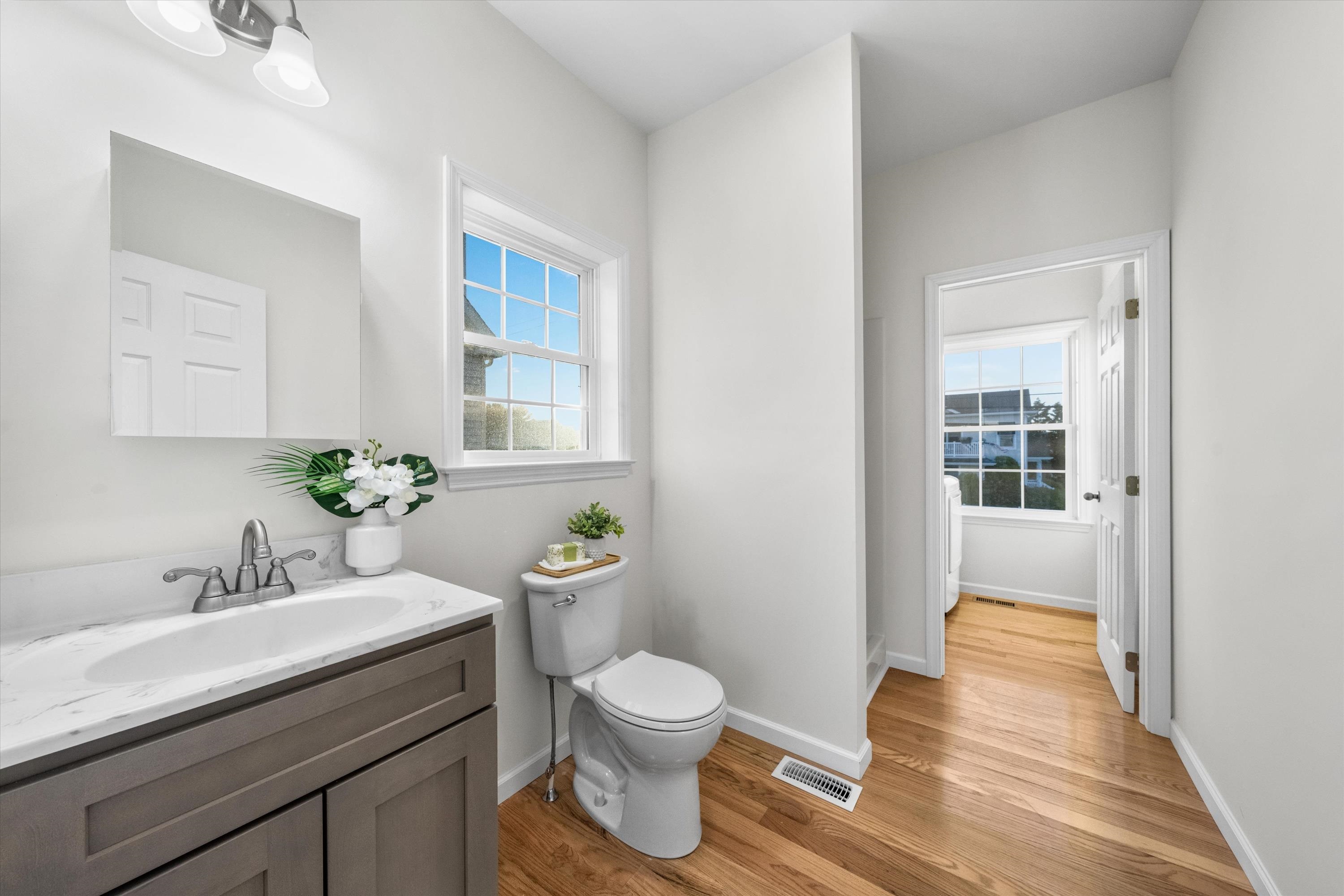 Bright bathroom with a gray vanity, white countertop, toilet, and hardwood floors.