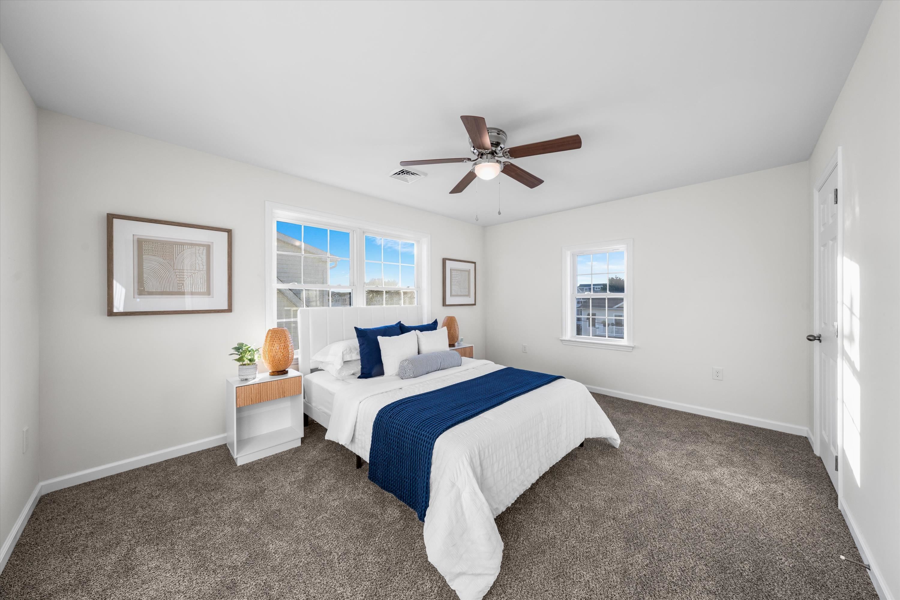 Staged bedroom with a white bed, navy throw, gray carpet, and ceiling fan.