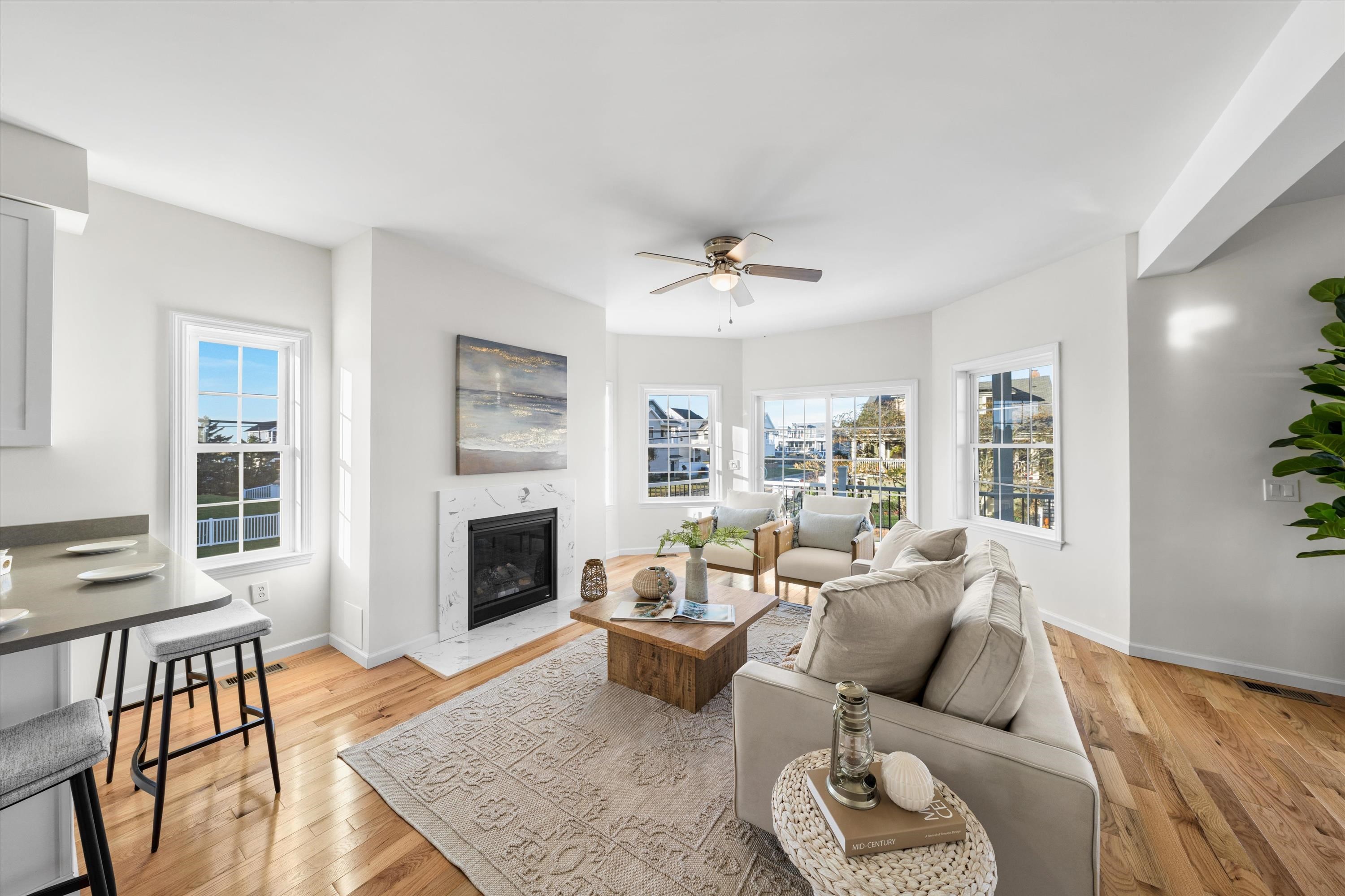 Bright living room with a marble fireplace, wood floors, neutral seating, and large windows overlooking a marina.