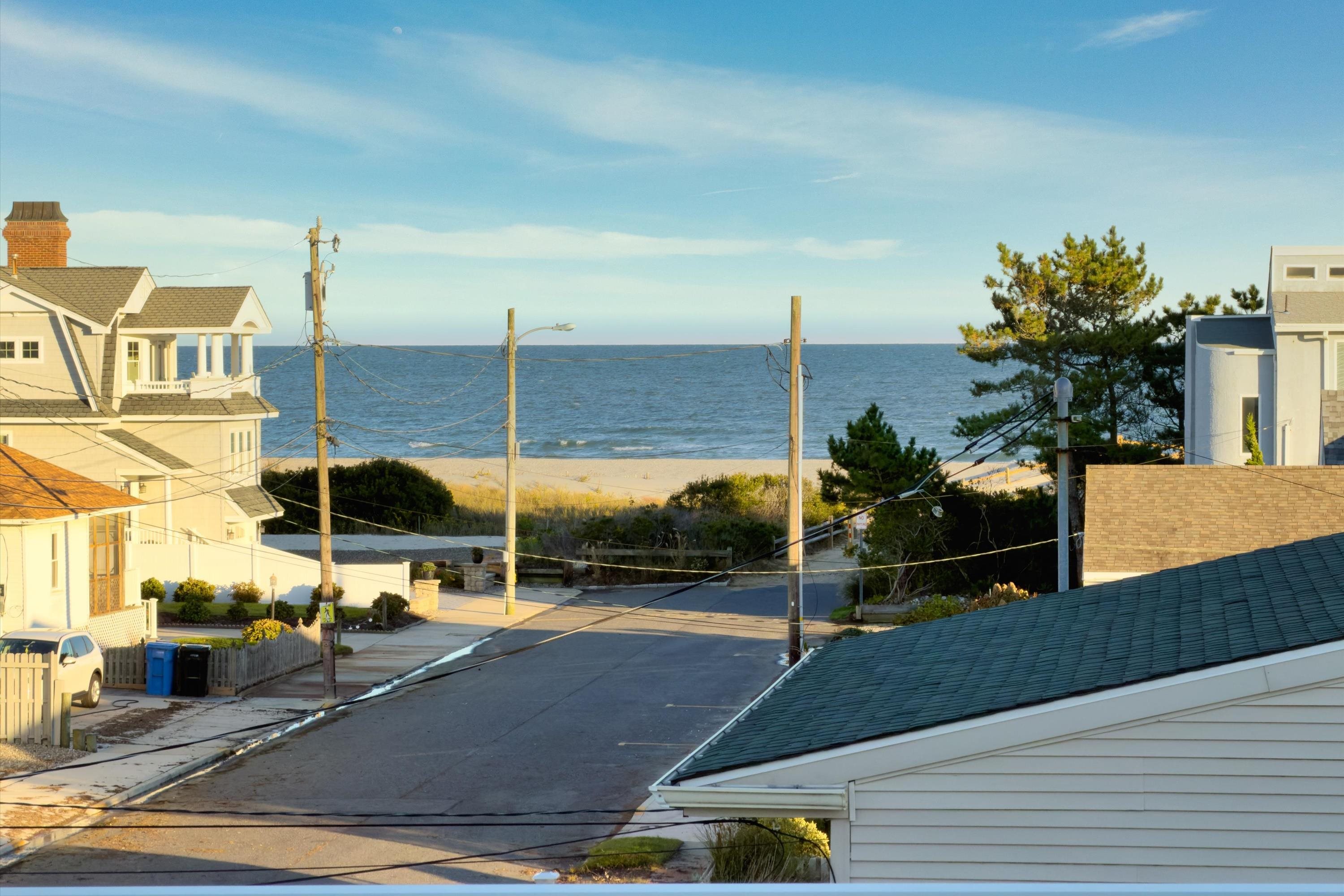 Ocean and sandy beach beyond a coastal street, framed by neighboring homes and utility lines.