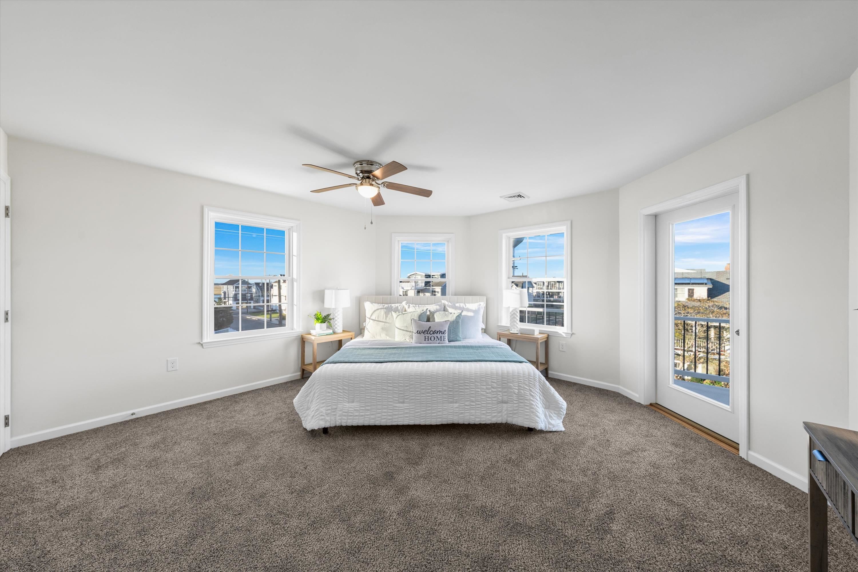 Furnished bedroom with a centered bed, gray carpet, and windows overlooking neighboring homes.