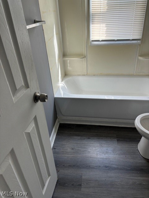 Bathroom with a white bathtub beneath a window and dark wood-look flooring.