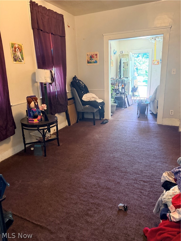 Carpeted living room with purple curtains and a wide doorway into a bright adjoining room.