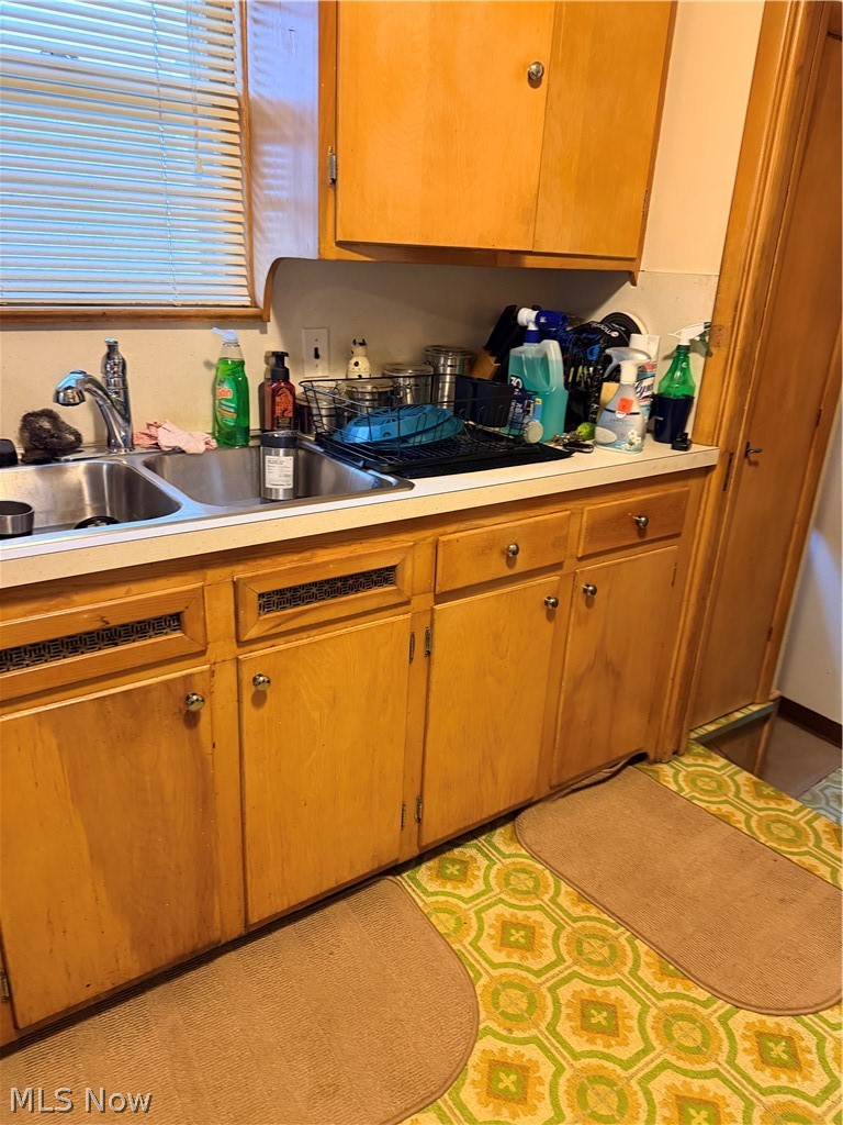 Kitchen sink and crowded counter beneath honey-colored wood cabinets, with a green geometric floor covering.