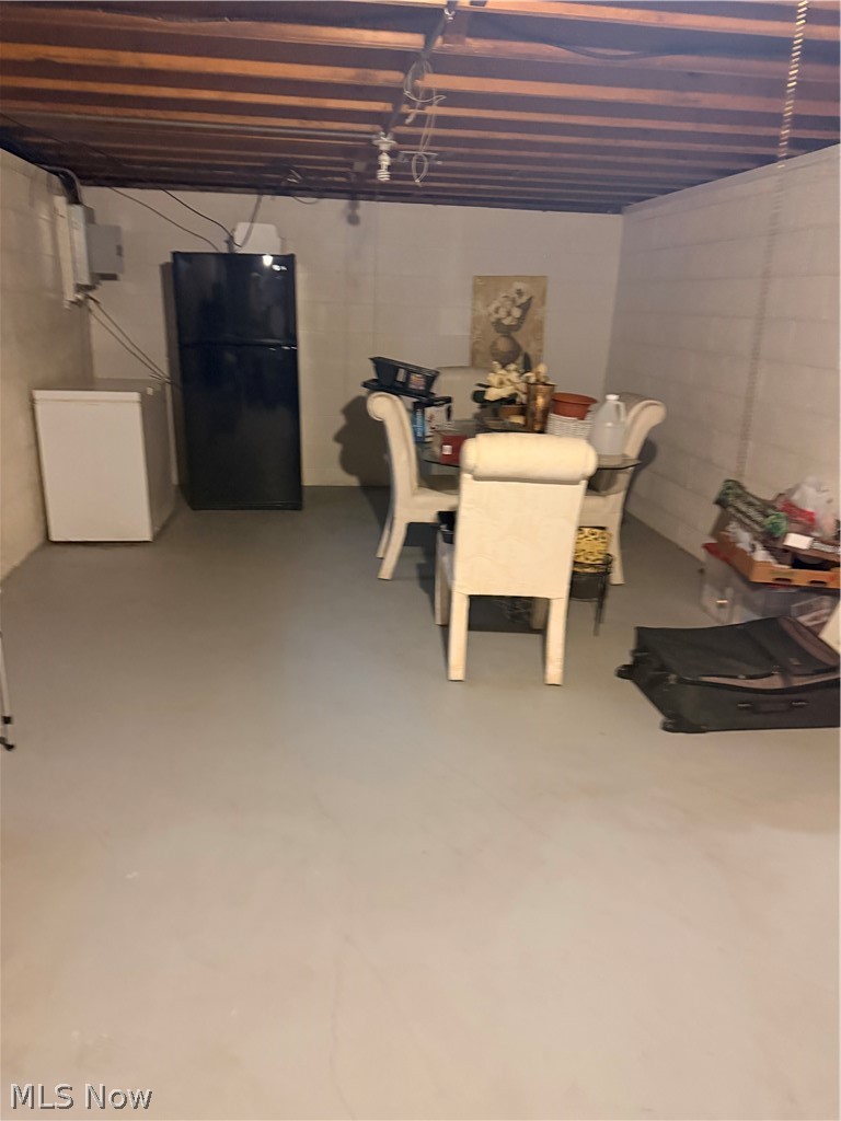 Basement room with exposed ceiling joists, block walls, and a dining table beside a refrigerator.