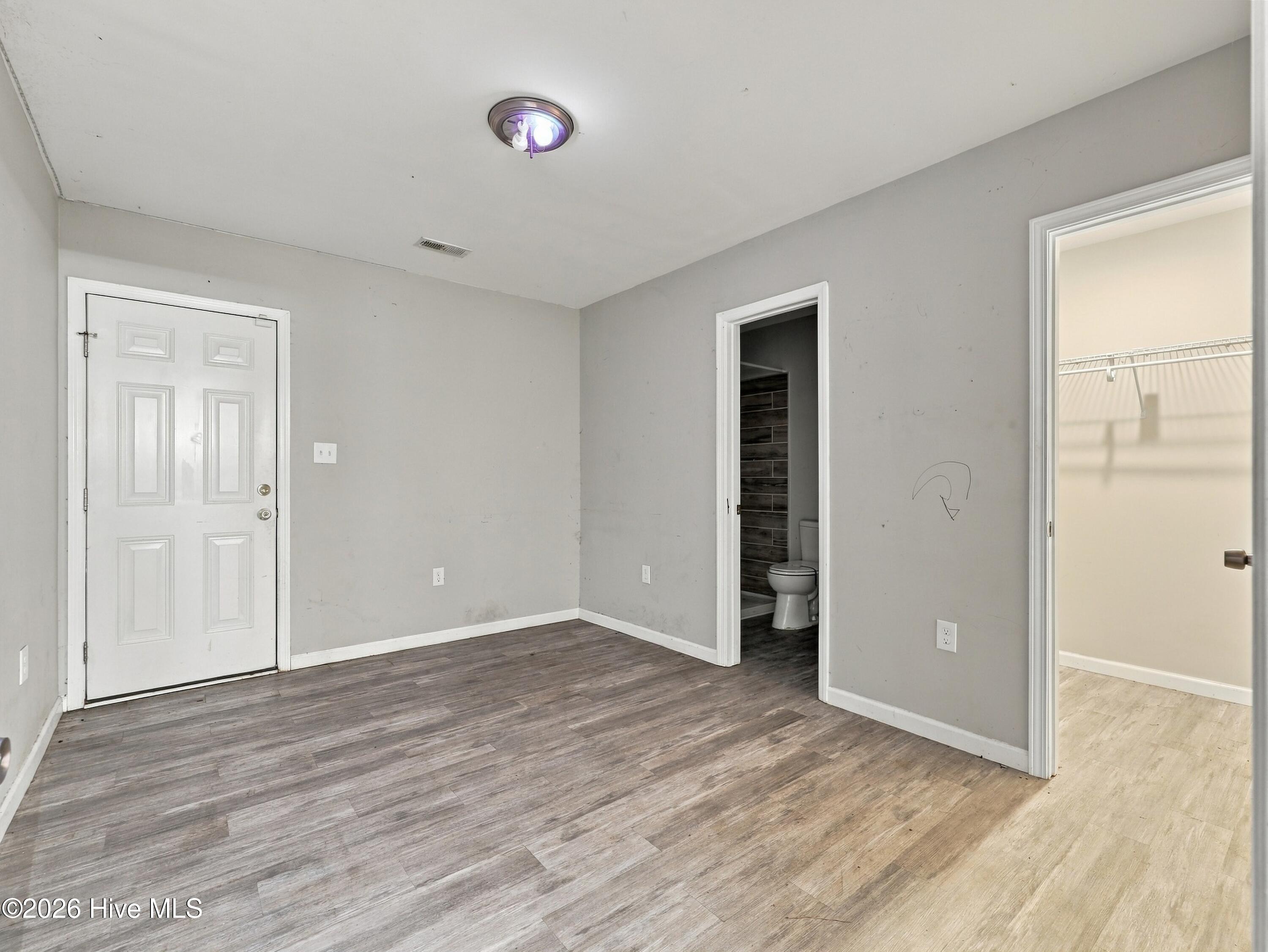 Unfurnished room with plank flooring, a front door, and open bathroom and closet doorways.