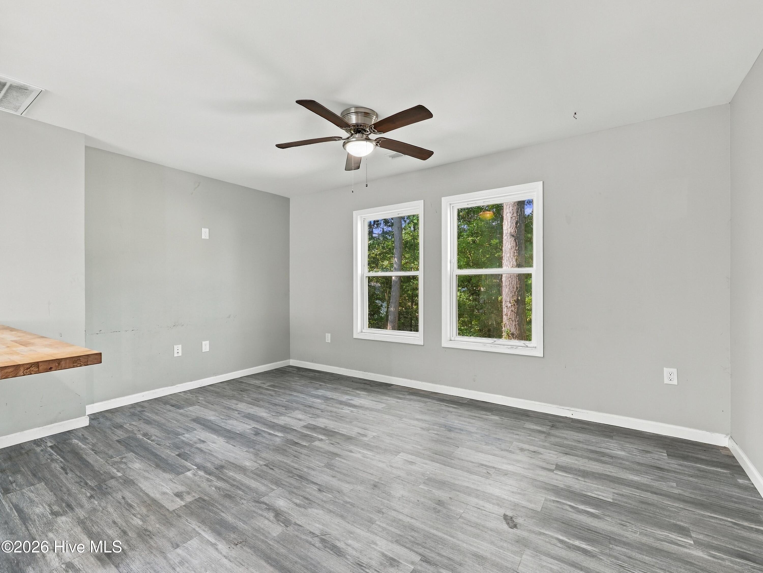 Empty room with gray plank flooring, two tree-facing windows, and a ceiling fan.