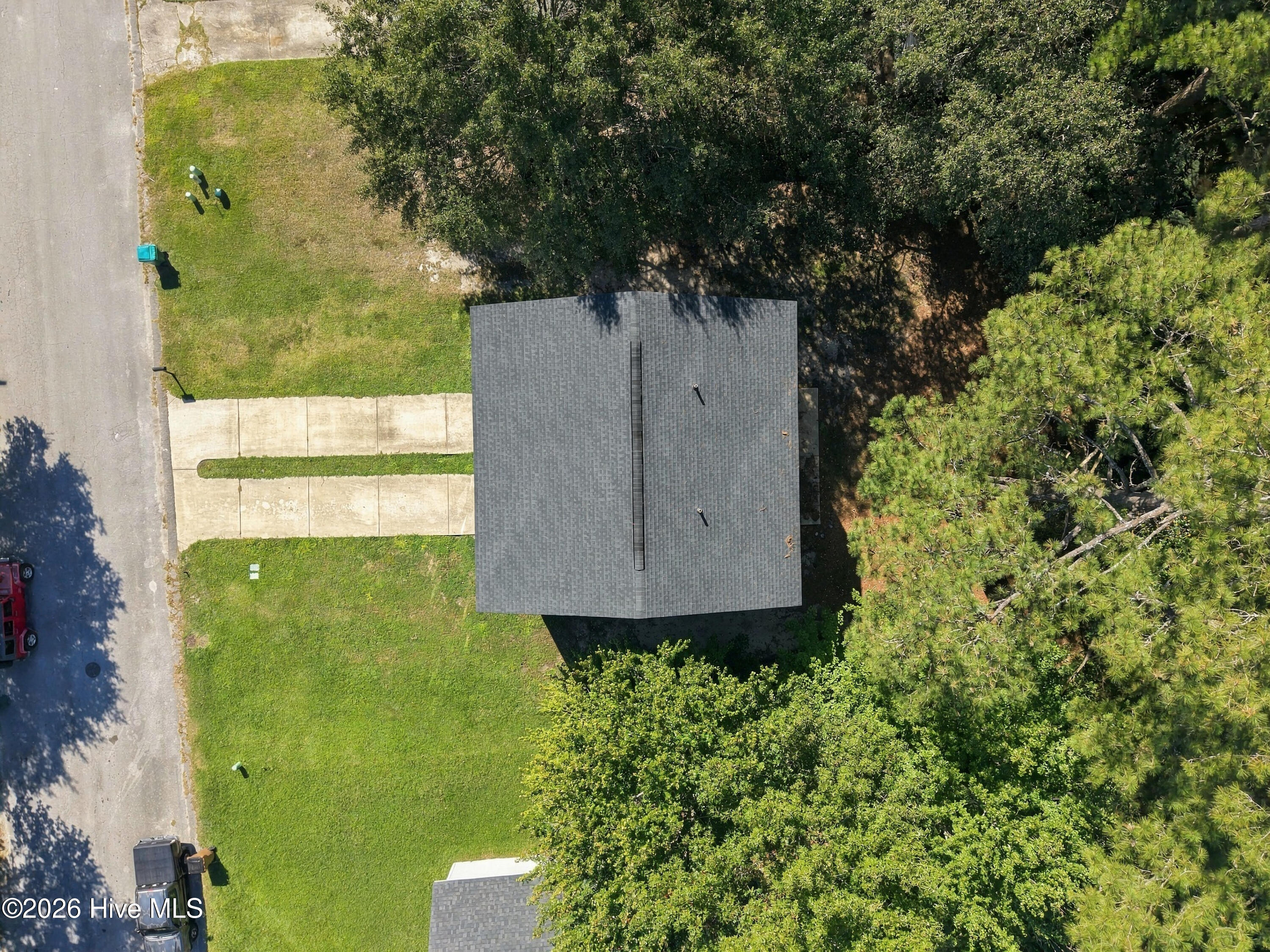 Aerial view of a gray-roofed house, concrete driveway, lawn, and surrounding mature trees.