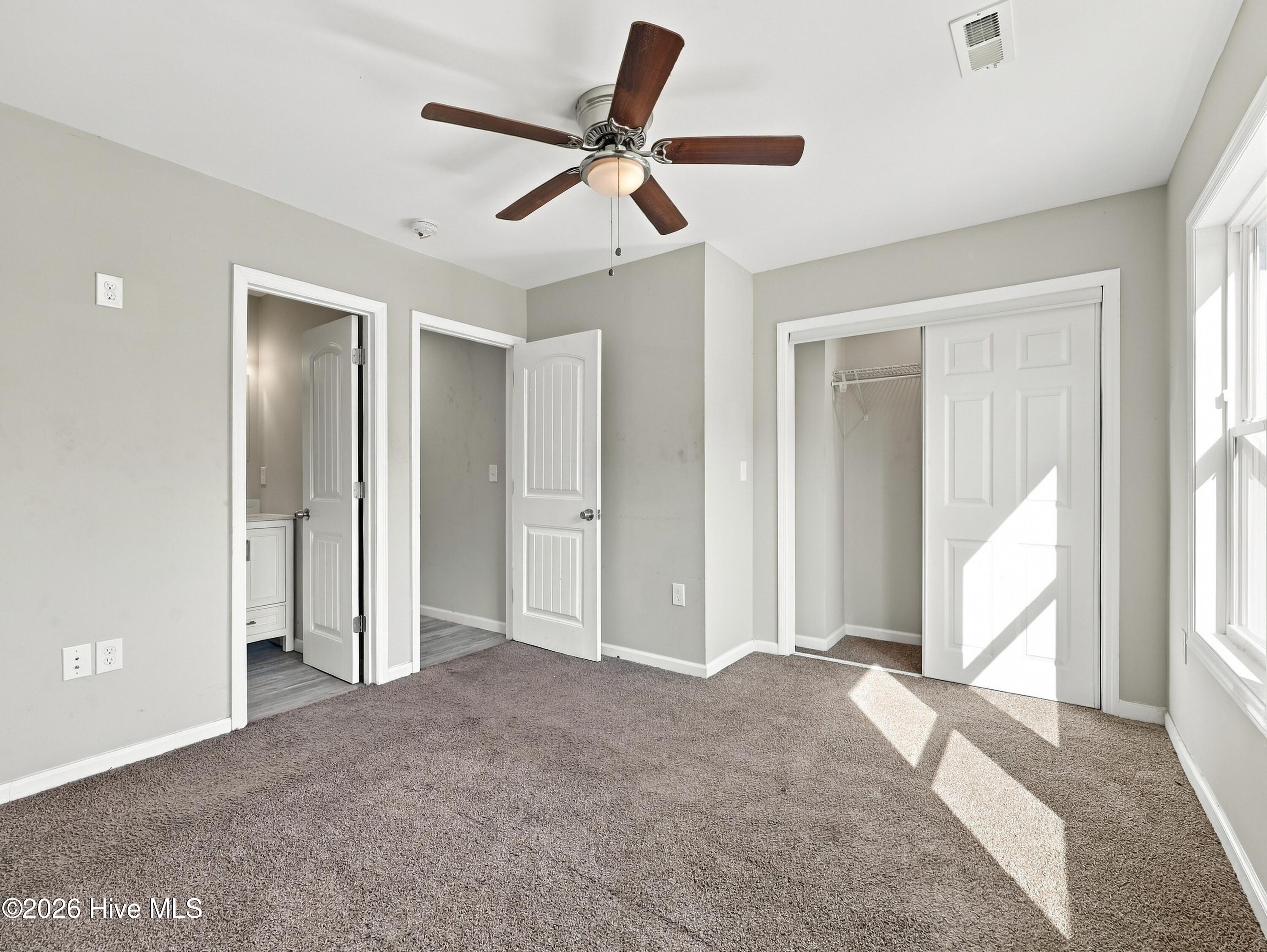 Empty carpeted bedroom with a ceiling fan, sliding closet doors, and sunlight streaming through windows.